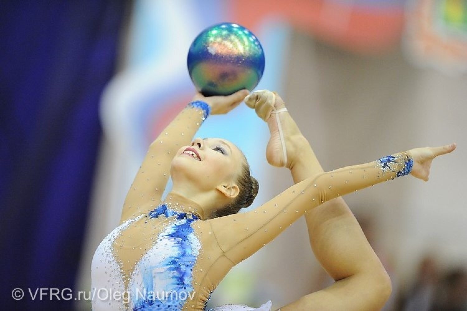 A young female rhythmic gymnast in a light blue and white leotard with rhinestones performs a routine with a colorful ball, stretching her arms and smiling.