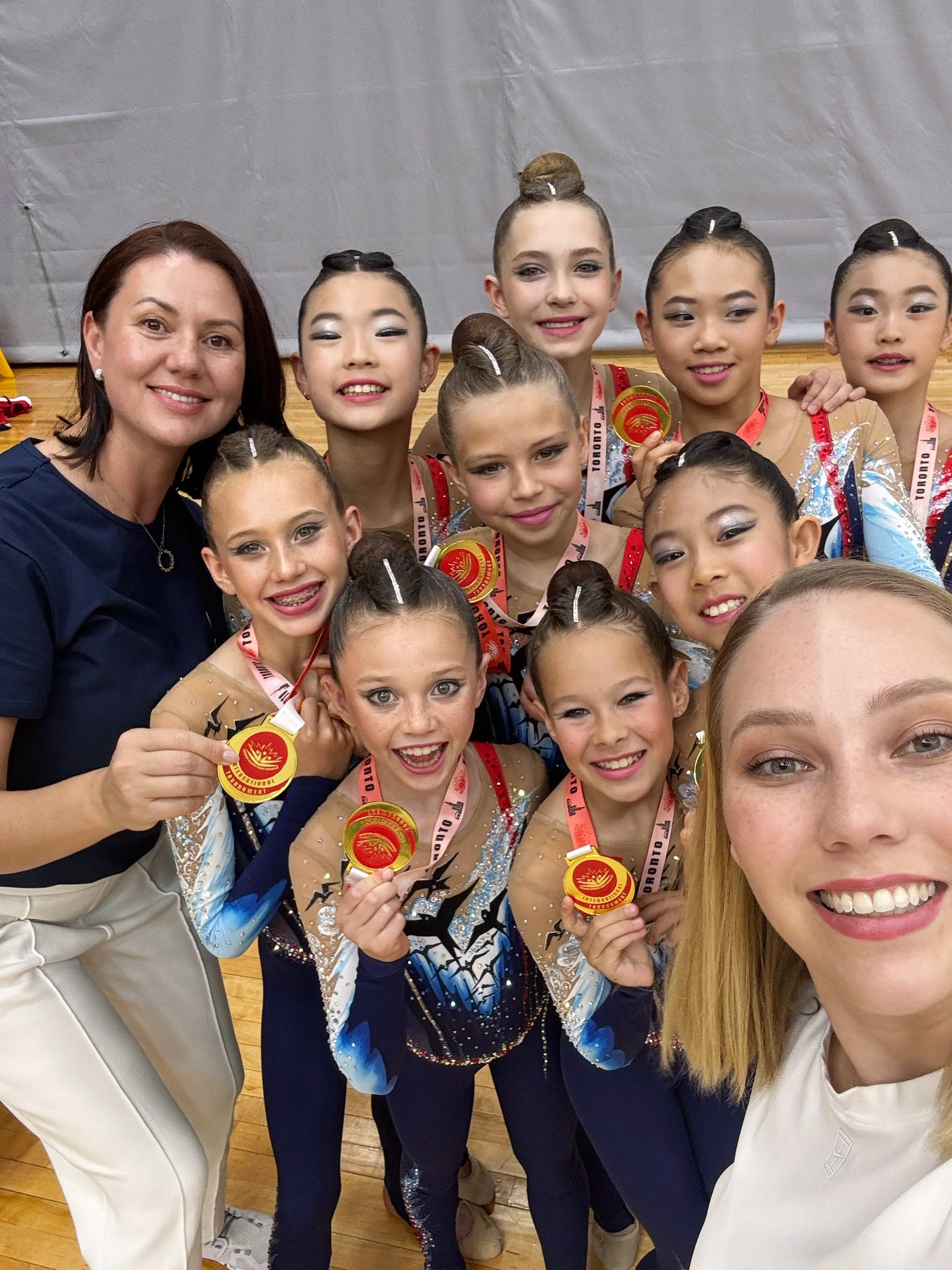 A group of young female gymnasts in colorful leotards holding medals, posing with two adult women, likely coaches, after a competition.
