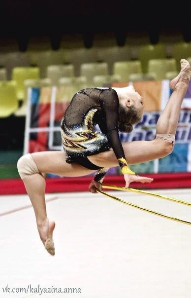 A young female rhythmic gymnast performing a jump with a split and back bend, wearing a black and gold leotard, inside a sports arena.