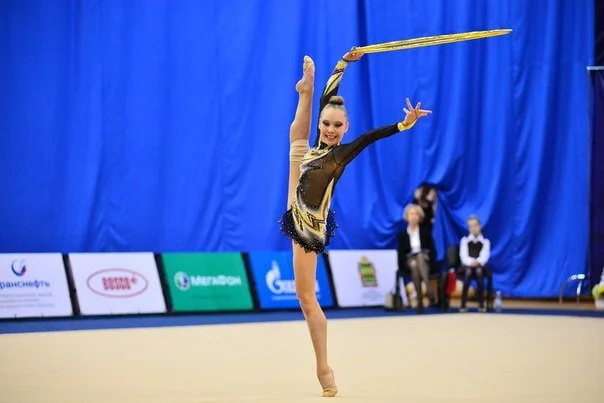 A female rhythmic gymnast performing a move with a golden stick during a competition, with a blue background and judges or officials seated in the background.