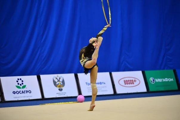 A gymnast performing an acrobatic move on a gym floor with a blue backdrop and sponsor banners in the background.