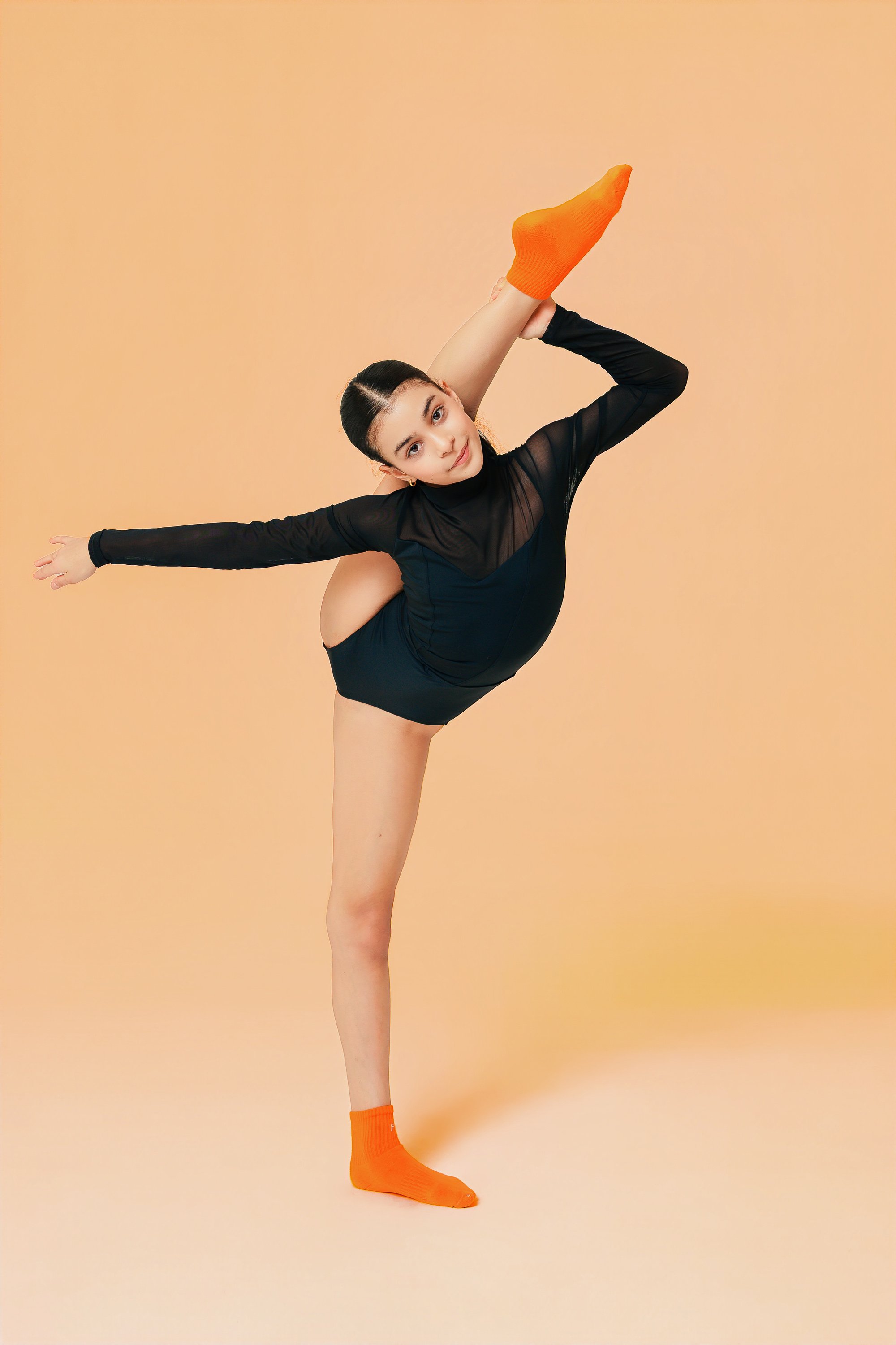 A young gymnast, in black dance attire, shows how flexible she can be by performing a standing split, one leg raised high as she balances on the other.