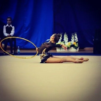 A rhythmic gymnast performing a routine on the floor with a hula hoop, with a judge or coach sitting in the background.