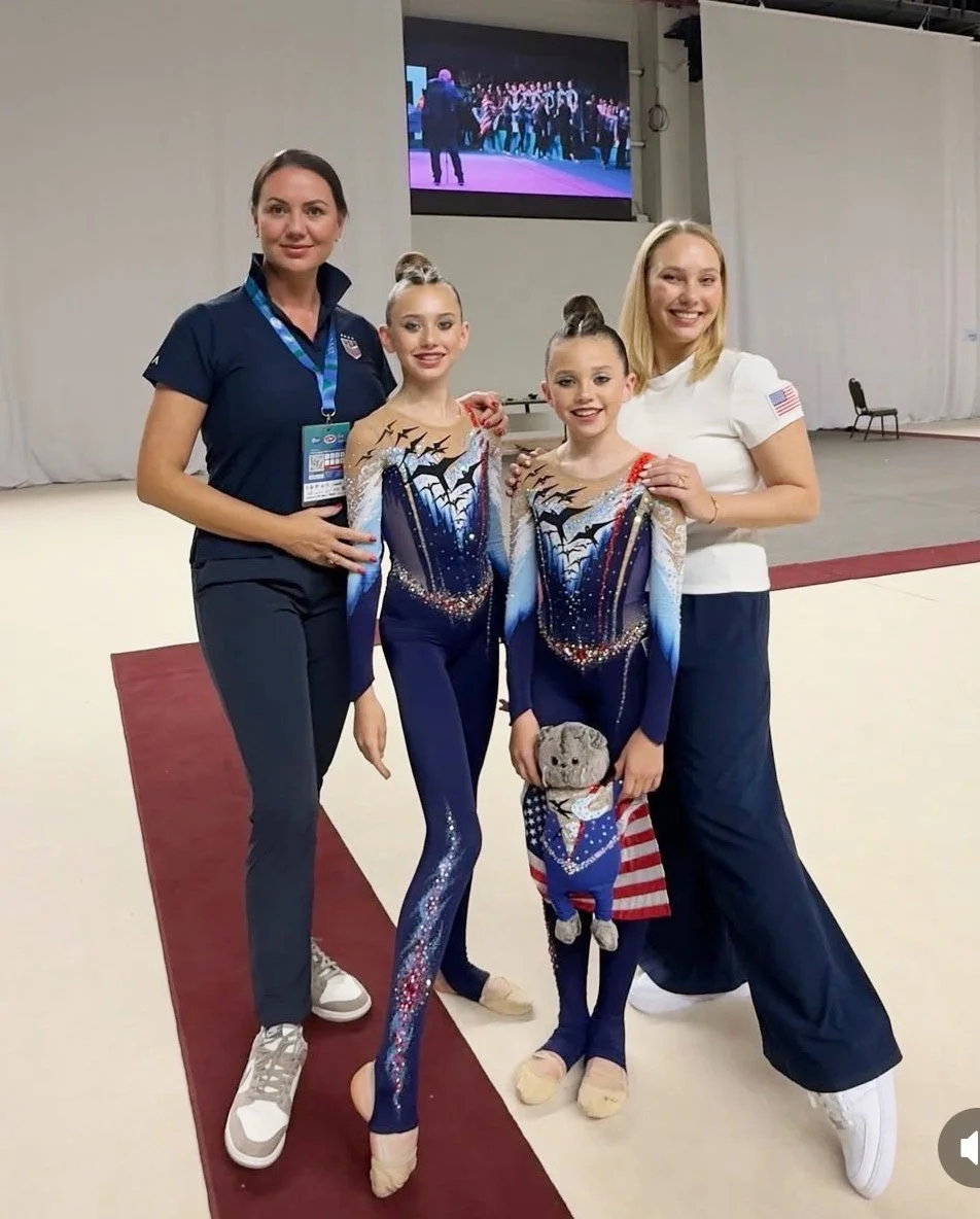 Two adult women and two young girls posing together backstage at a gymnastics event, with a large TV screen showing a group of people in the background.