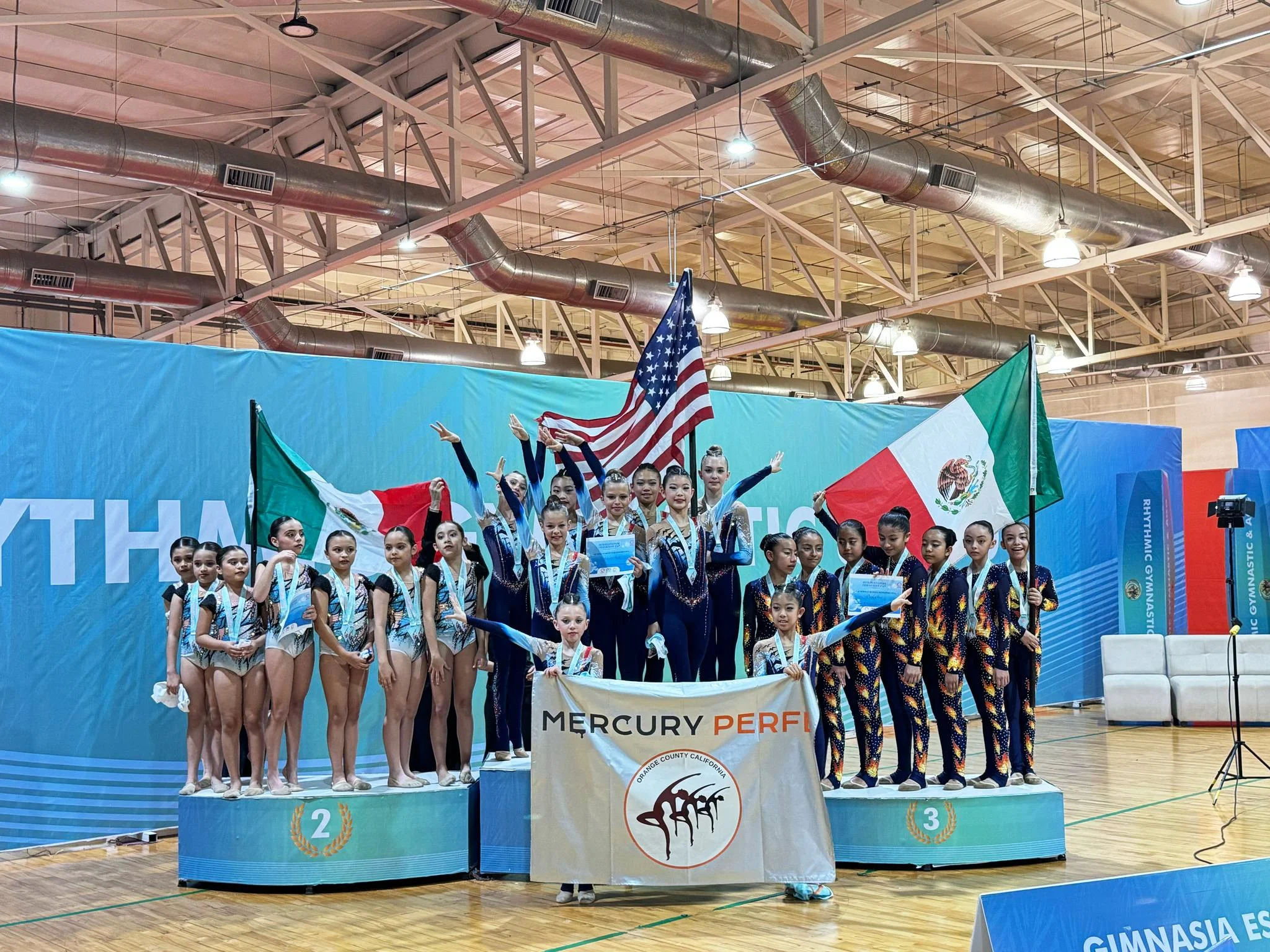 Group of young female gymnasts on the winners podium at a gymnastics competition, holding flags of the United States and Mexico, with medals around their necks and a banner reading 'Mercury Perfe' in front.