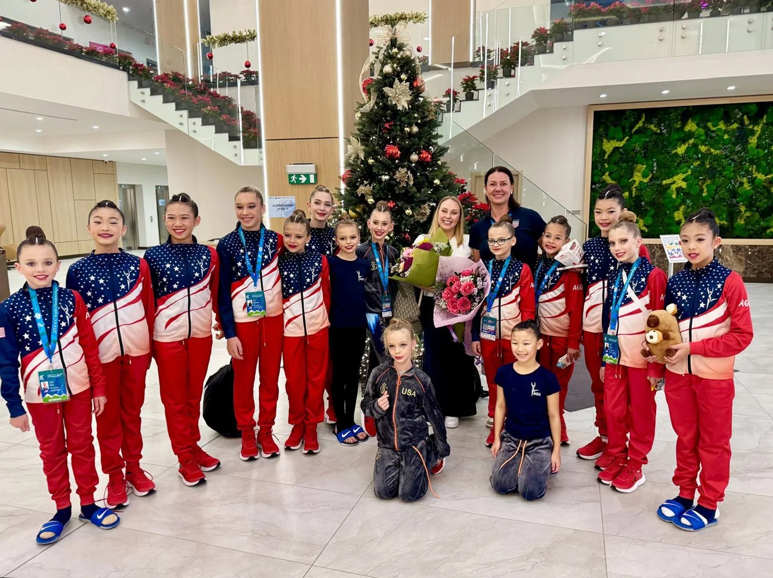 Group of young female gymnasts in red, white, and blue uniforms posing for a photo in front of a decorated Christmas tree in an indoor setting with modern design elements.