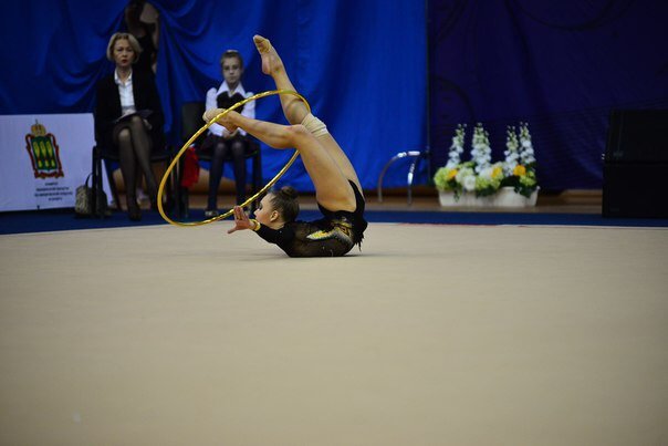 A rhythmic gymnast performing a move with a yellow hoop on a beige carpeted floor, with judges or audience members seated in the background.