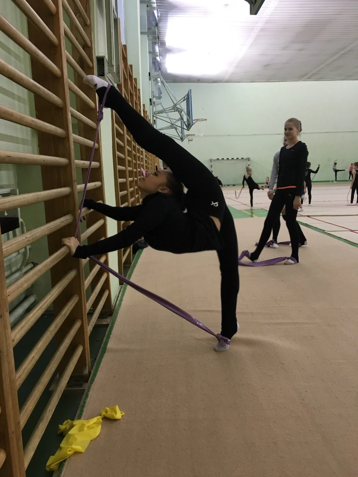 Gymnast practicing a stretch on the wall bars while holding resistance bands, with other gymnasts stretching in the background.