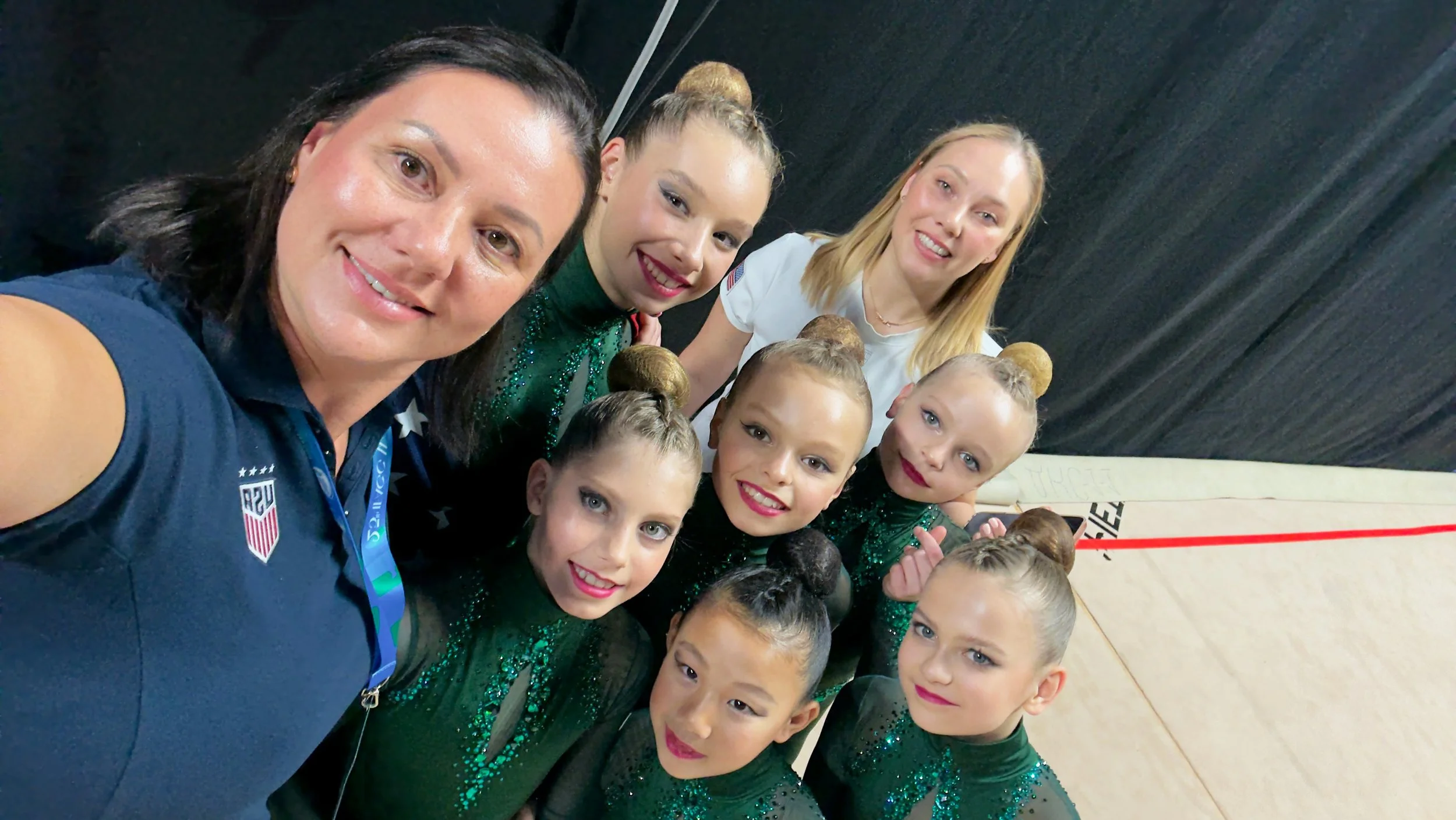 Group of young female gymnasts in green leotards with bun hairstyles, taking a selfie with their coaches backstage.