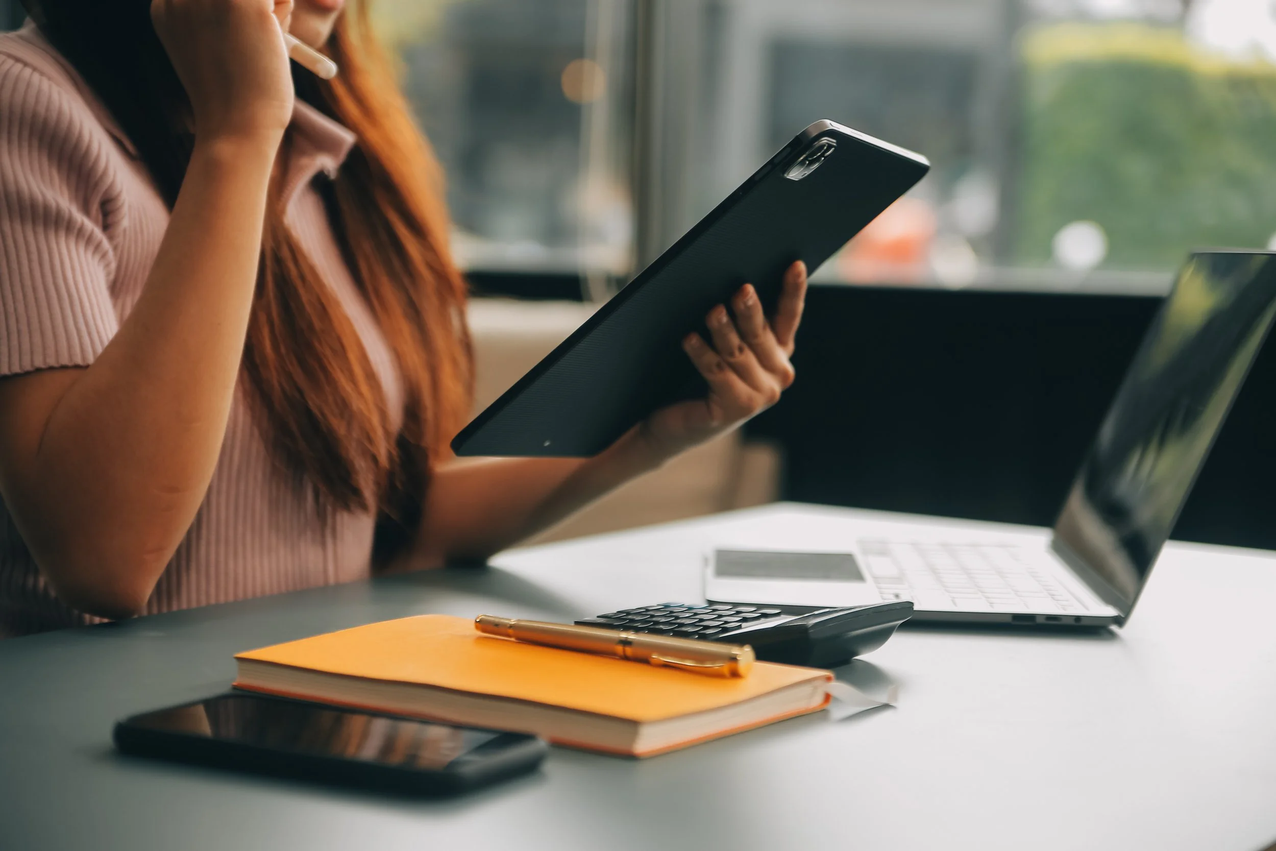 A woman with long red hair is sitting at a table with a laptop, a smartphone, a notebook, and a pen, holding a tablet in her right hand and touching her face with her left hand.