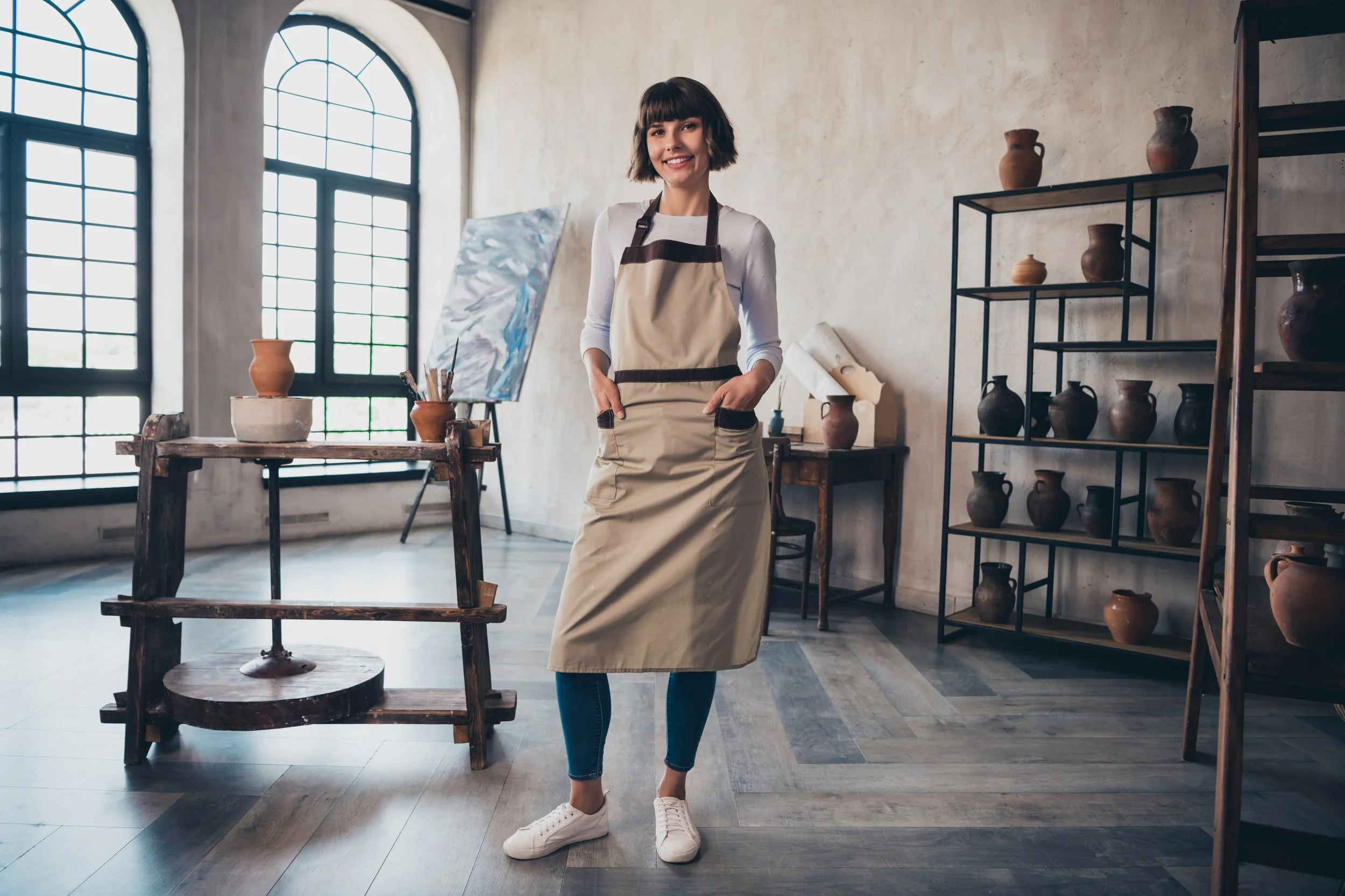 A woman standing inside an art studio, wearing an apron, smiling with her hands in her pockets, surrounded by pottery and art supplies, with large windows in the background.