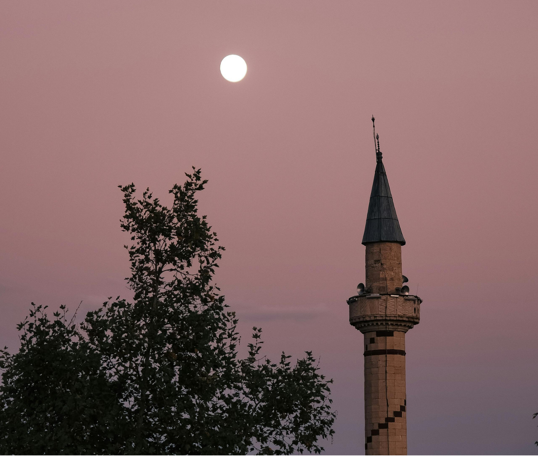 A mosque minaret with a pointed top and a small balcony, next to leafless trees, under a pinkish sky with a bright full moon.