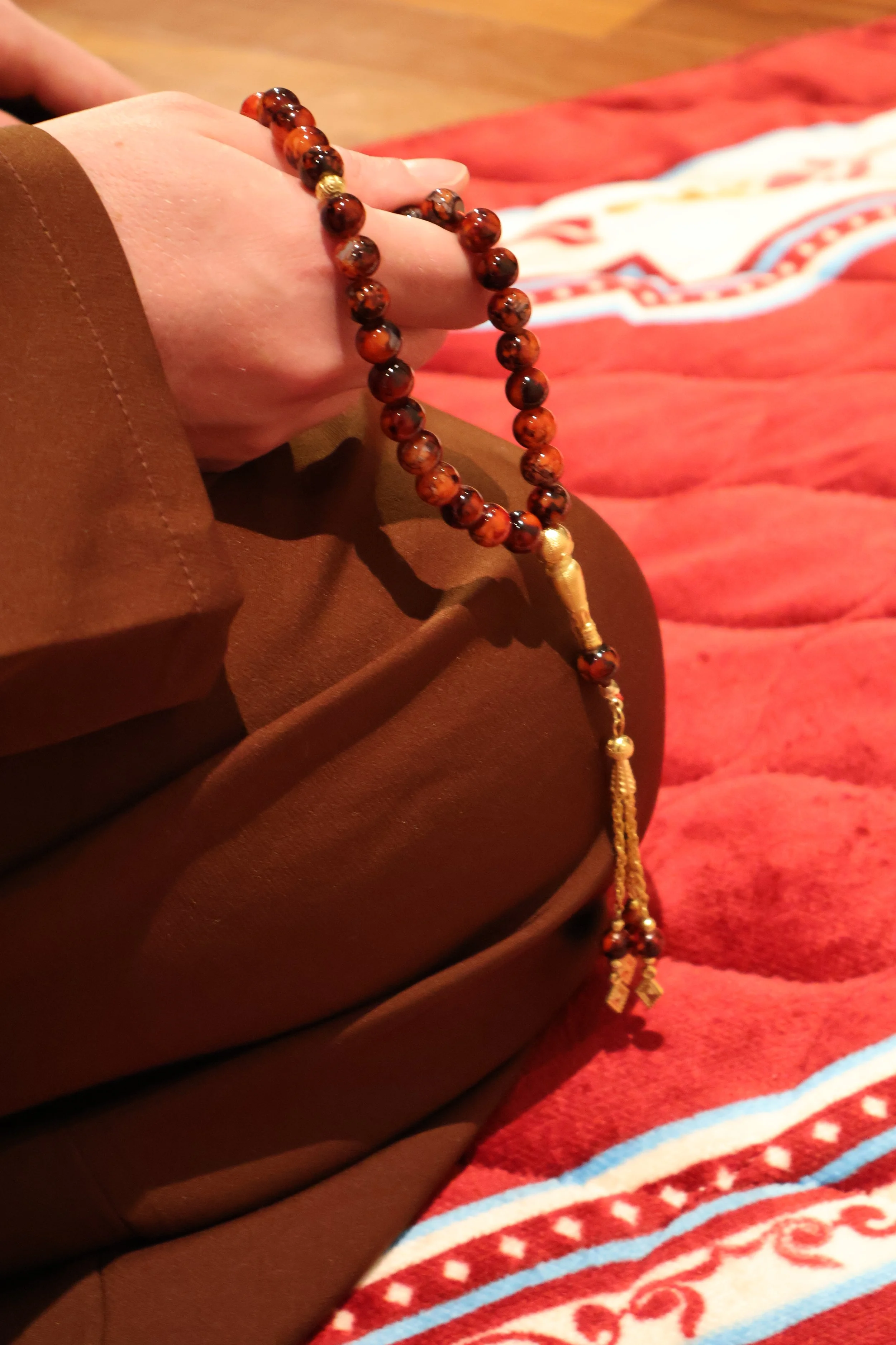 A person holding a string of prayer beads with brown and gold beads, with a red prayer mat in the background