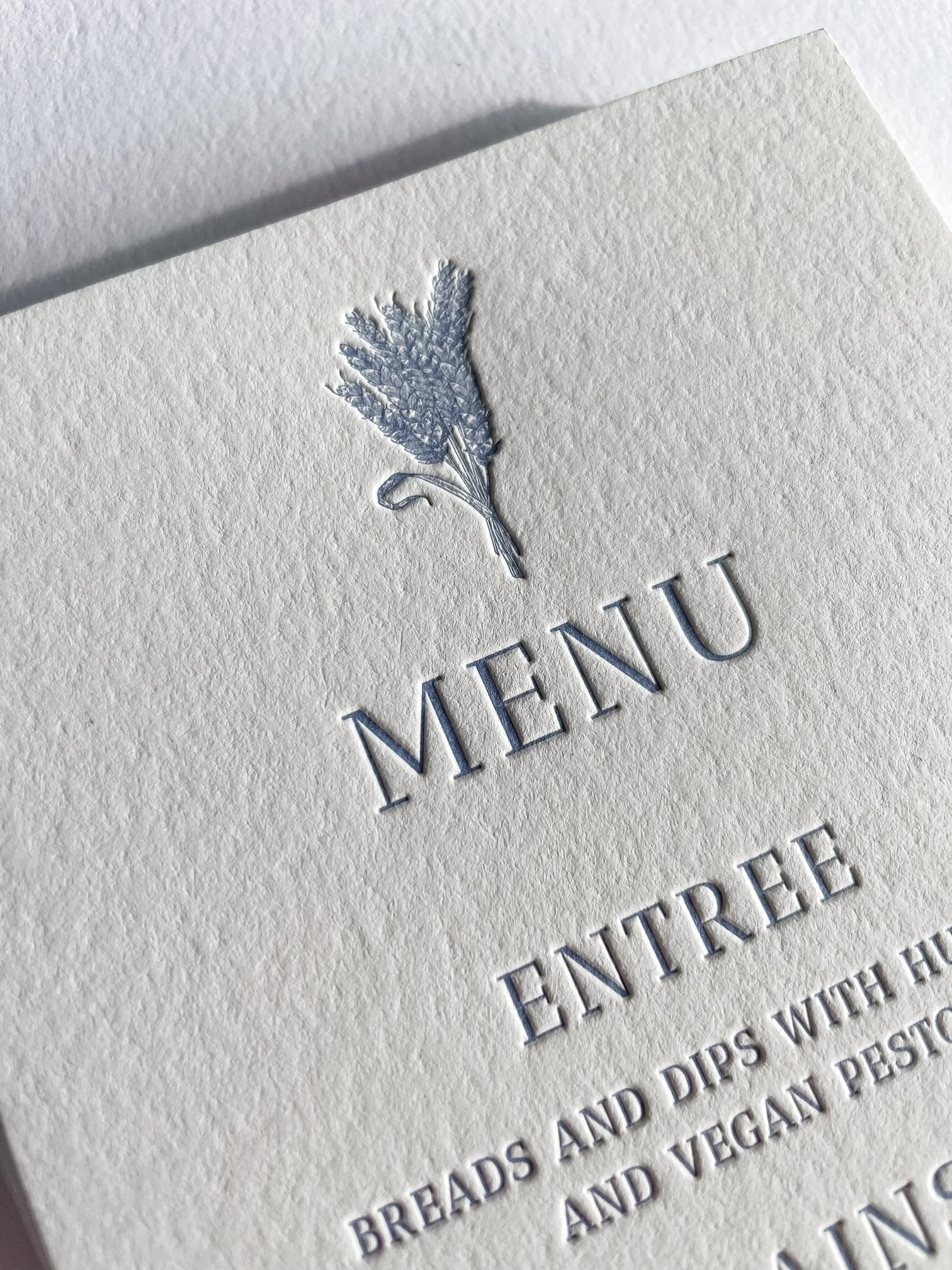 Close-up of a textured cream-colored paper with a silver embossed illustration of a sprig of lavender and printed text.