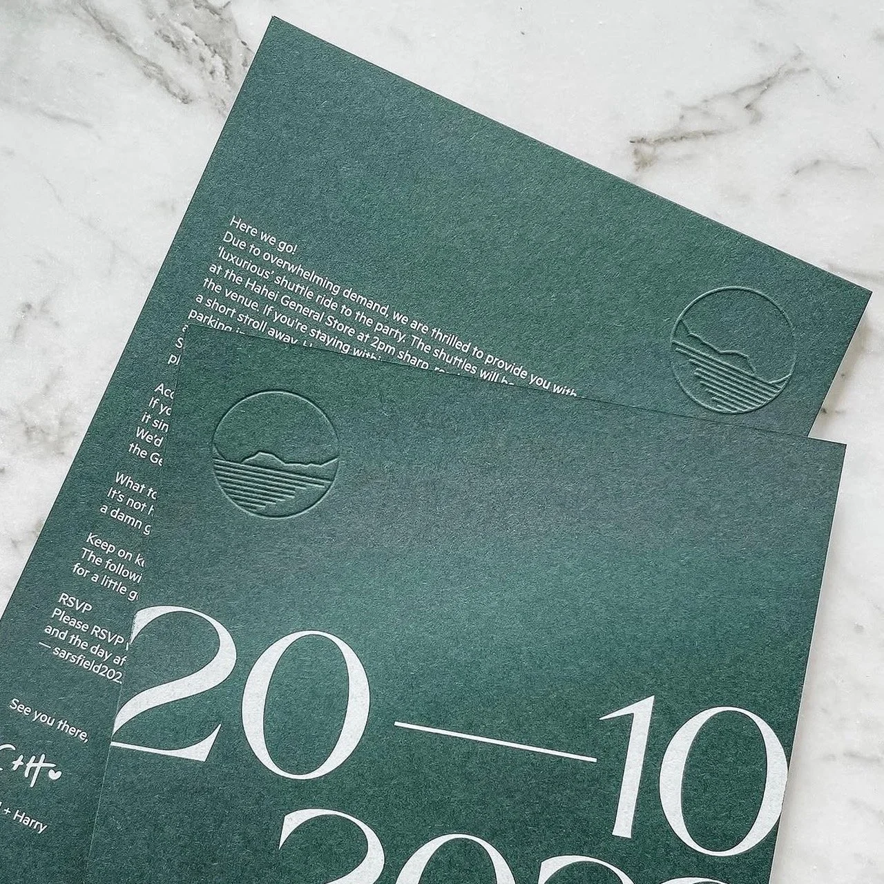 Close-up of dark green invitation or card with embossed logo of a sunset or mountain range over water, and white text with event details, on a light marble surface.