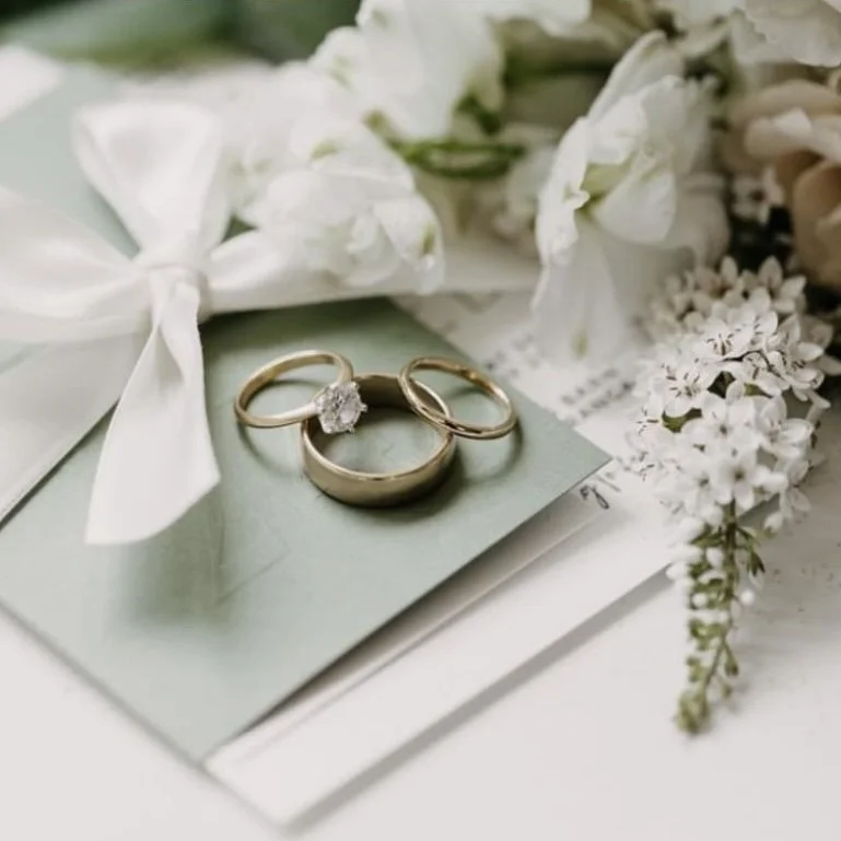 Three wedding rings placed on a light green card, with white flowers and a white ribbon tied in a bow nearby.