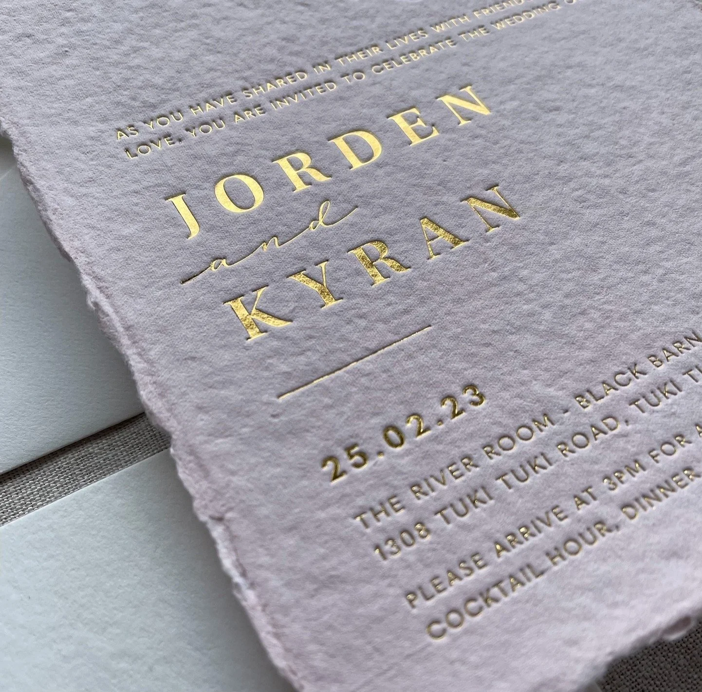Close-up of a wedding invitation with gold embossed text on textured paper, details about the event including date and location.