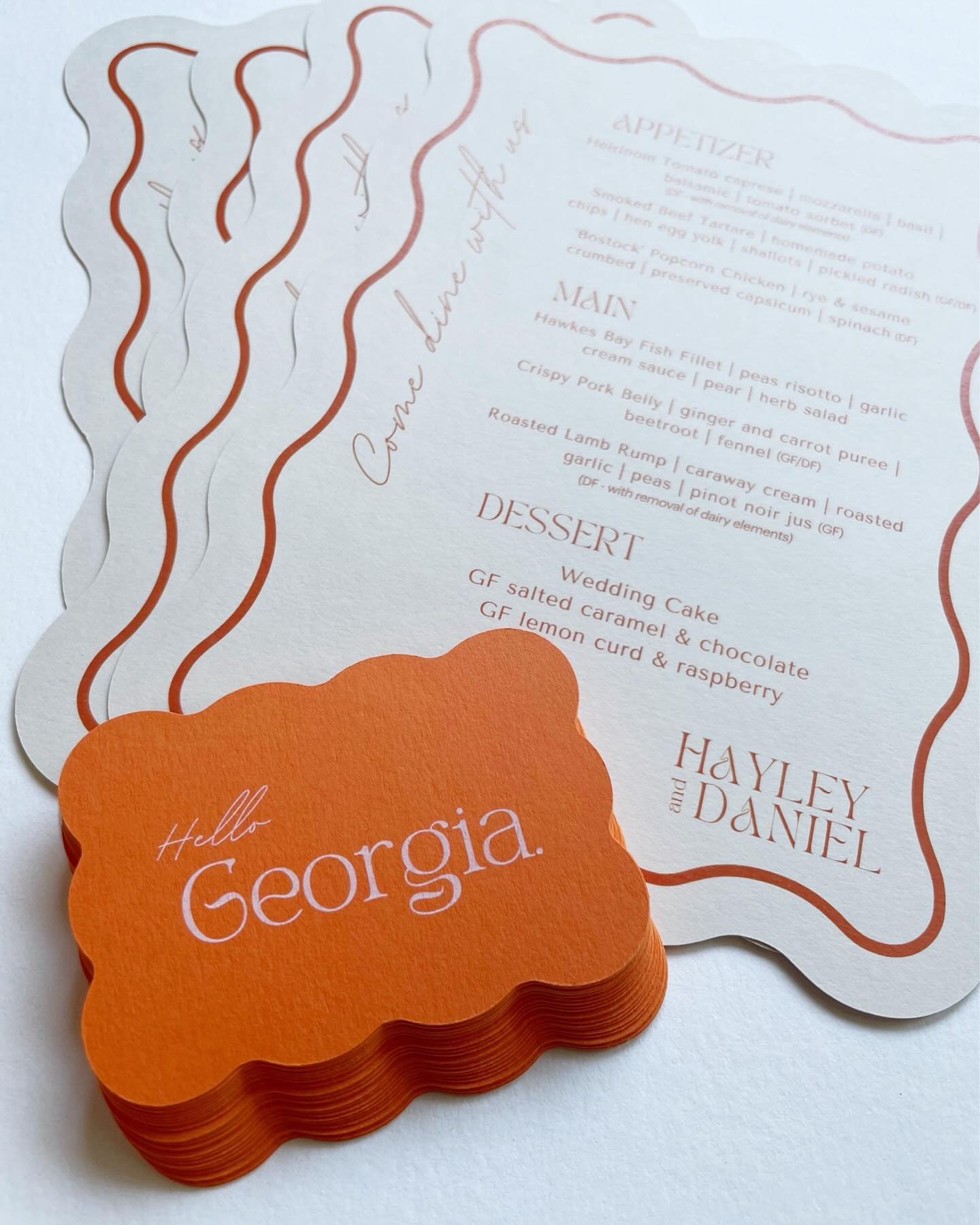 A stack of orange cards with the words 'Hello Georgia.' written on the top card, placed on a white surface with a decorative paper menu in the background.