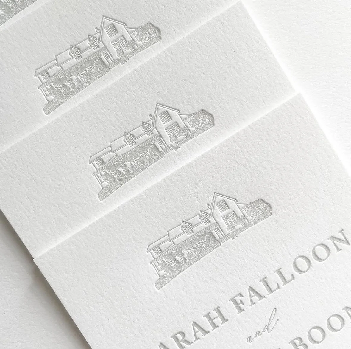 Close-up of white embossed wedding invitations with house and tree illustrations, and the text 'Sarah Fallon and...'