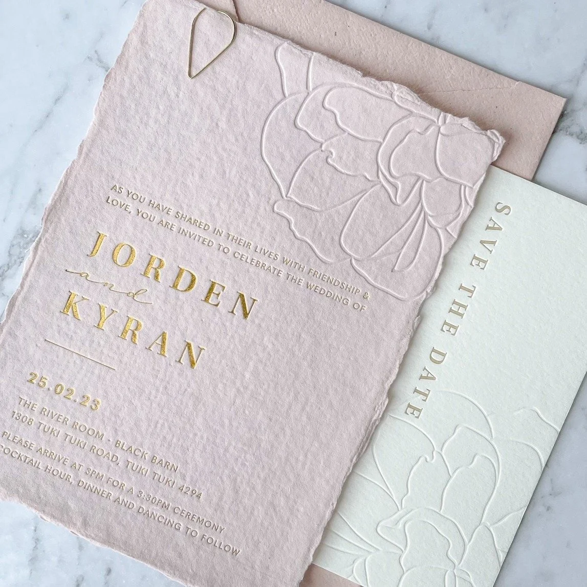 Wedding invitation card for Jorden and Kyran with gold embossed text on textured pink paper, including event details and floral embossed design, accompanied by matching RSVP card.