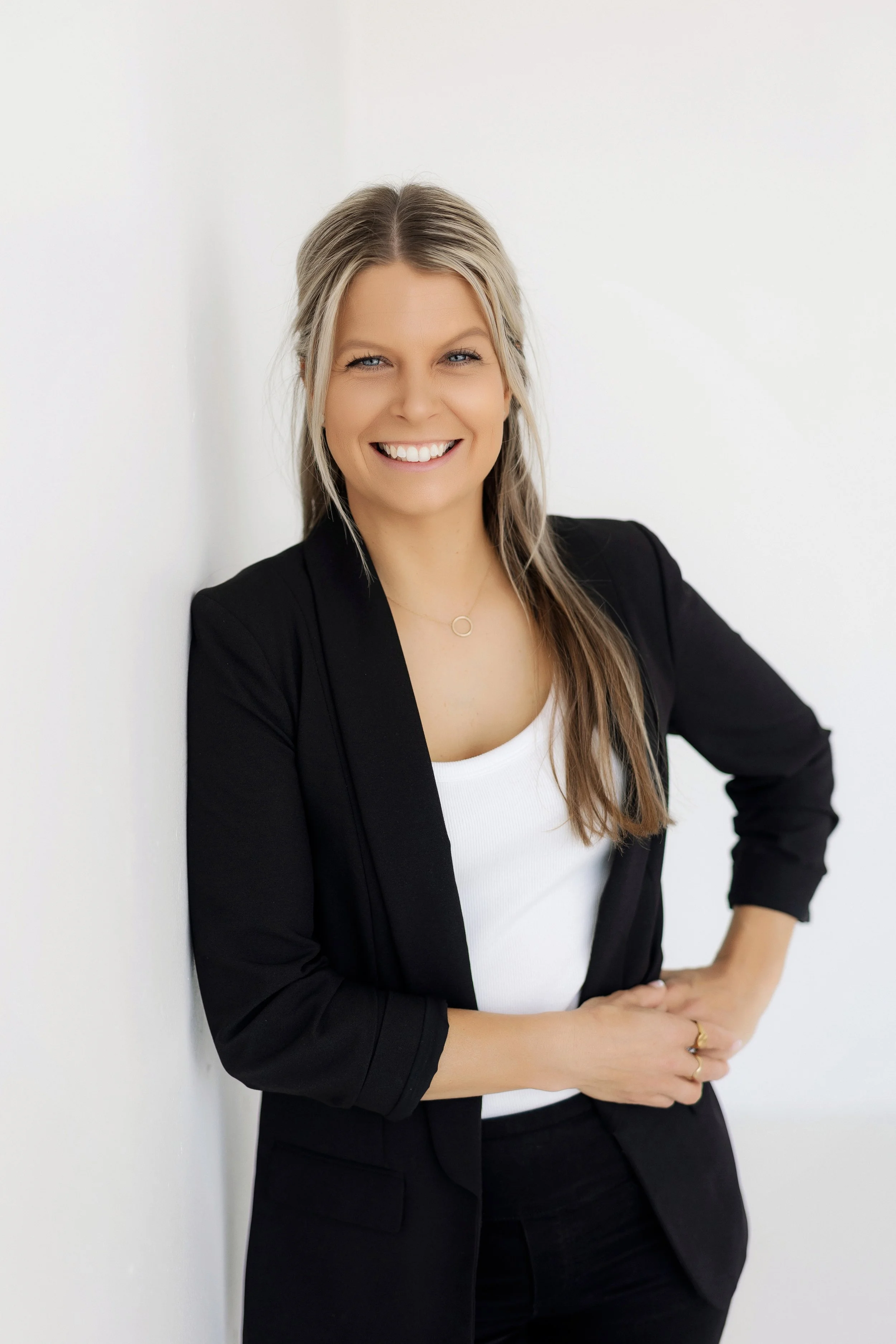 Woman in black blazer leaning on wall, one hand on hip and the other holding that hand, smiling.