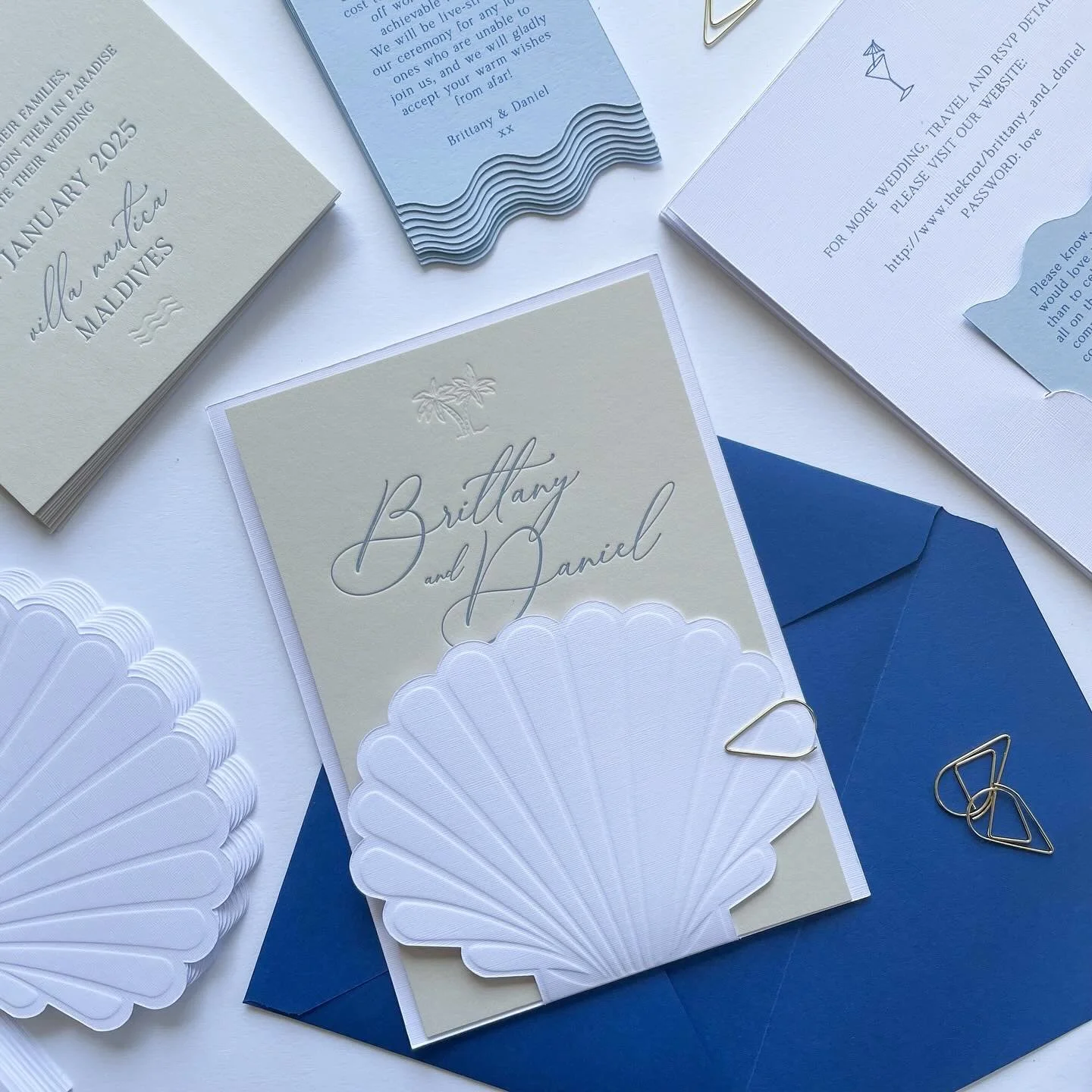 Wedding invitation cards with light blue, white, and navy blue colors, featuring decorative shells, palm trees, and elegant cursive text for Brittany and Daniel's wedding.