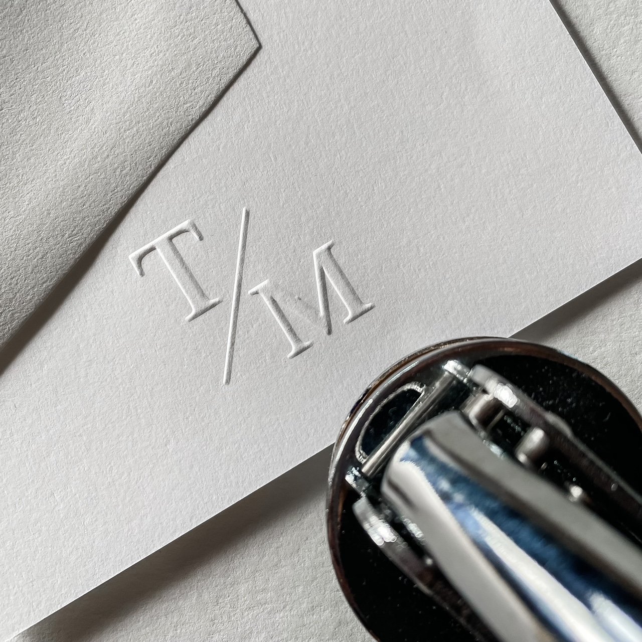 Edge of page with T & M initials with line in between embossed. Embosser at edge of frame.