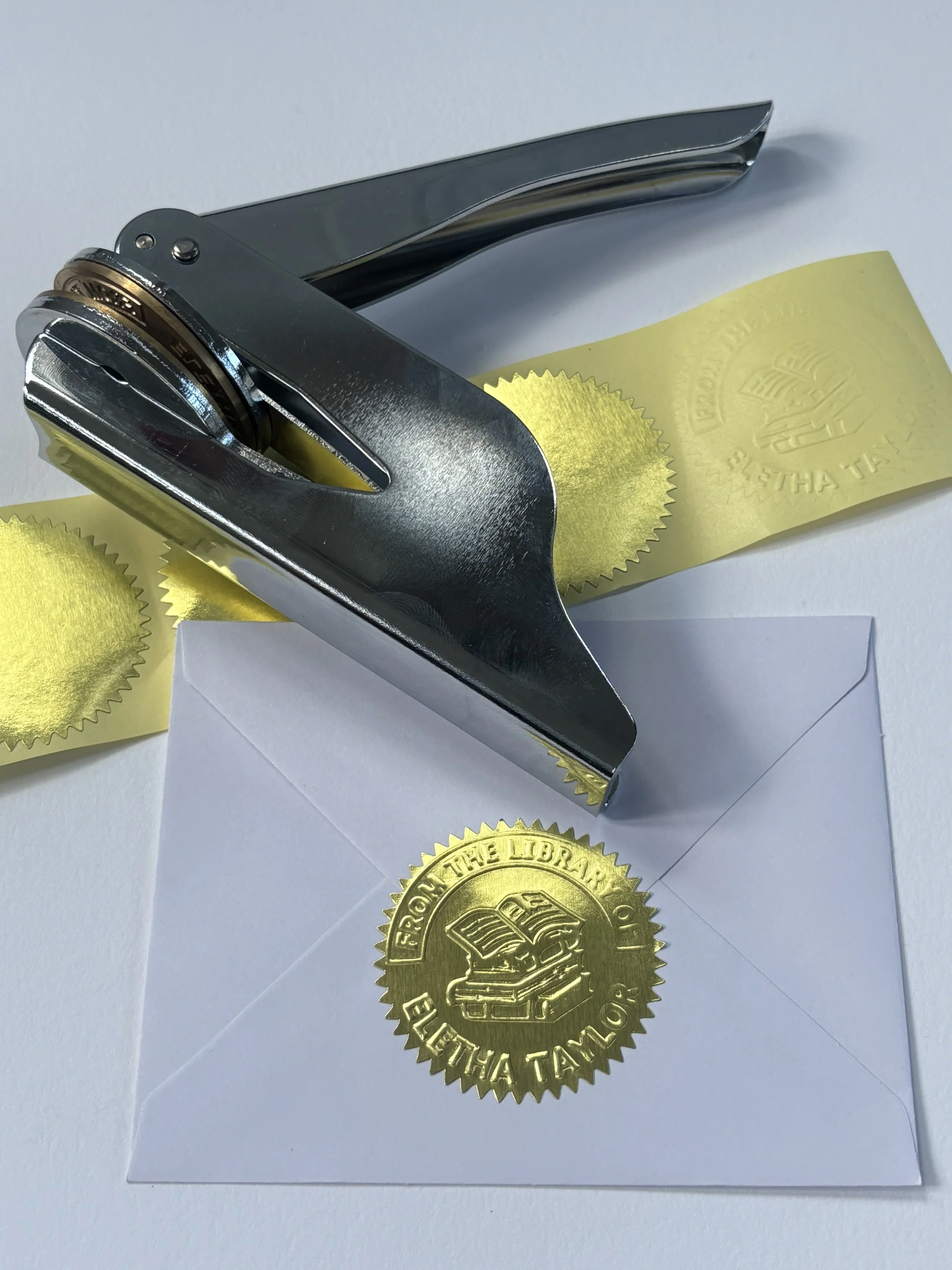 Silver embosser laying on top of blank gold foil stickers. Pressed gold foil sticker used to seal envelope.