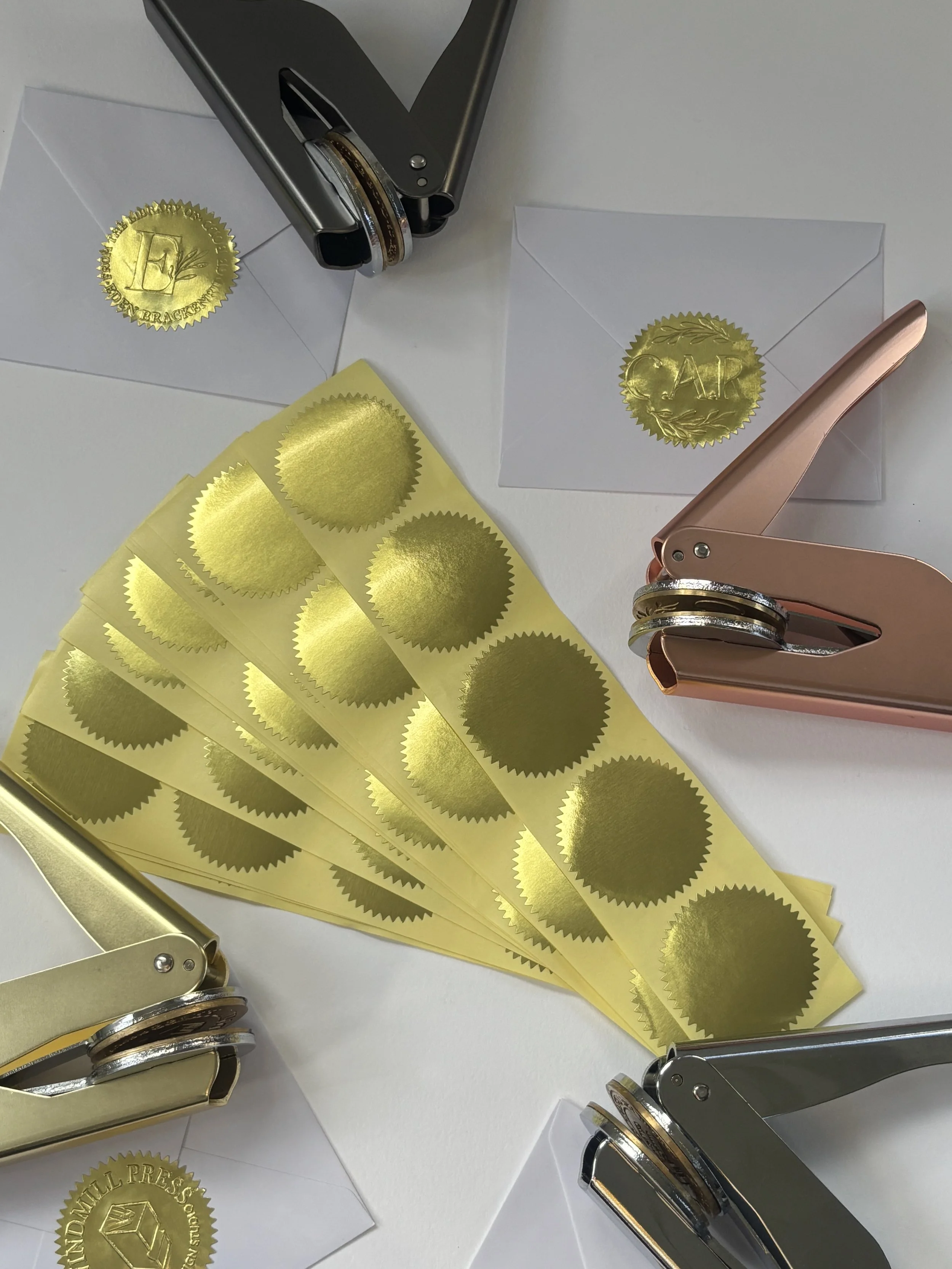 Overhead shot of four embossers laying amongst envelopes. Strips of five gold foil stickers laid in the centre.