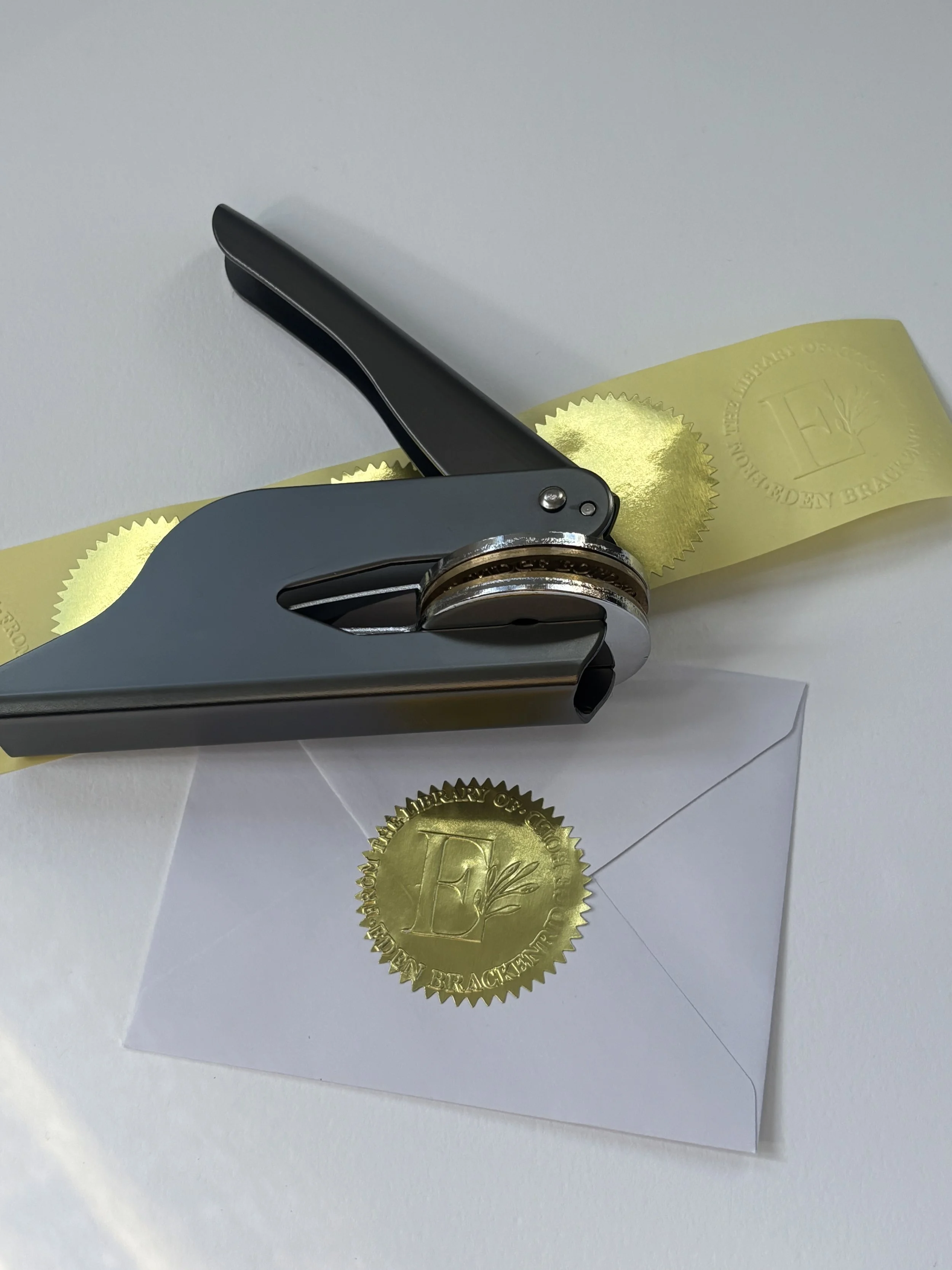 Gunmetal embosser laying on top of blank gold foil stickers. Pressed gold foil sticker used to seal envelope.