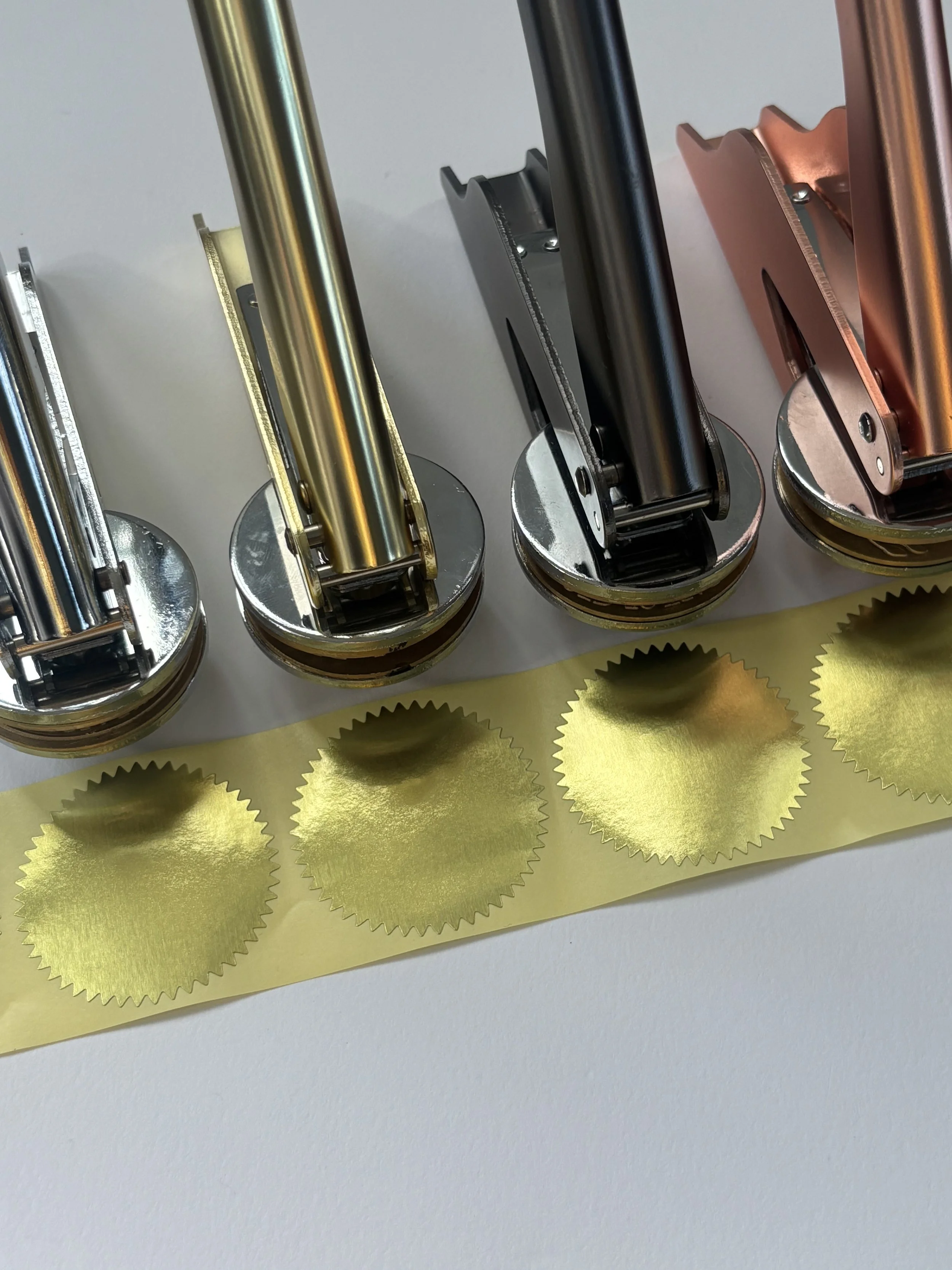 Embossers in various metals lined up in front of blank gold foil stickers.