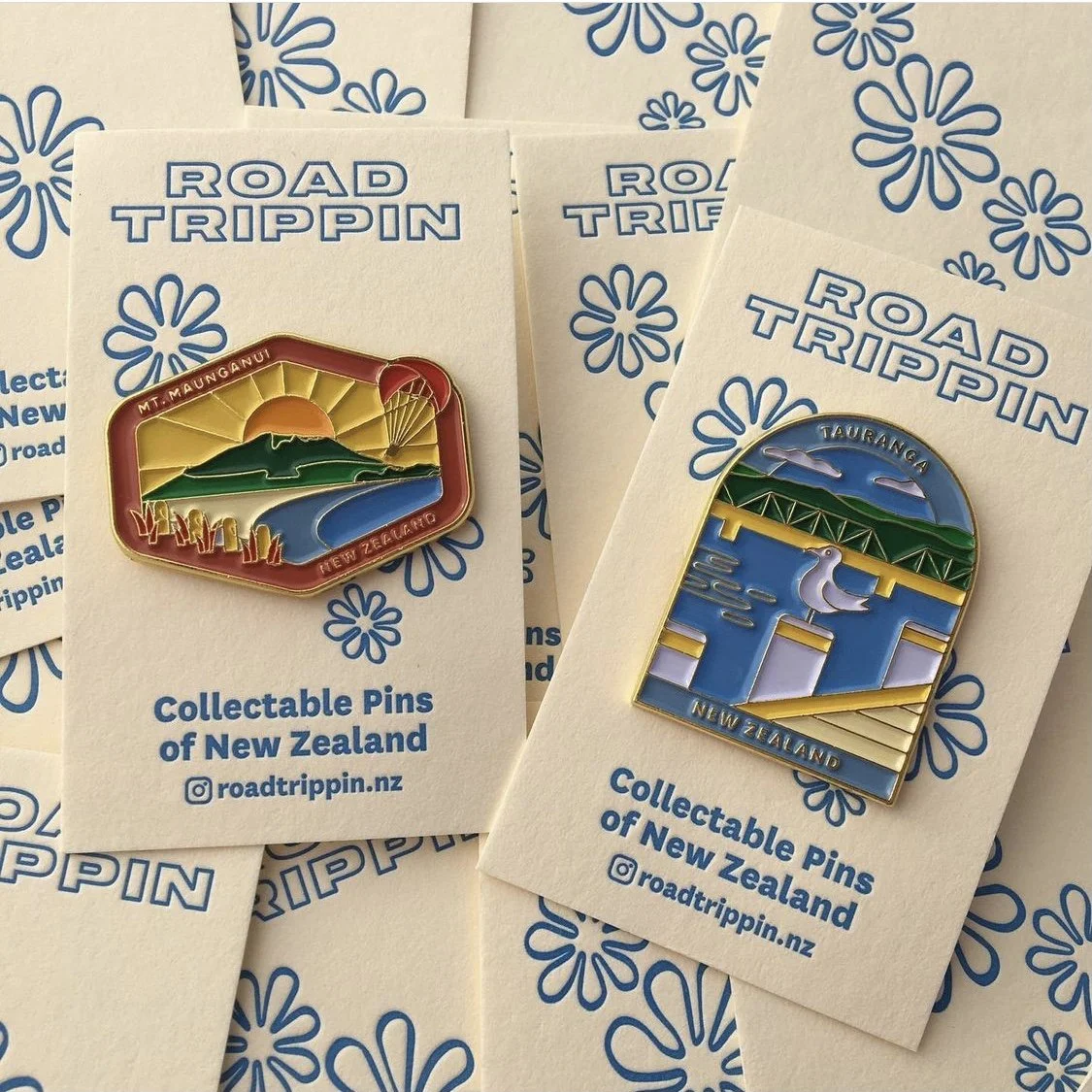 Two collectible pins from New Zealand, featuring landscape scenes, attached to branded cards with 'ROAD TRIPPIN' in blue at the top, and the Instagram handle '@roadtrippin.nz' at the bottom.