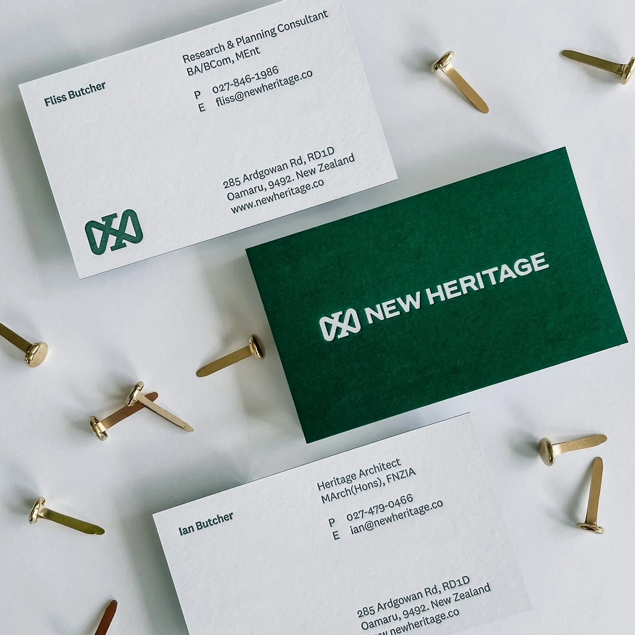 Business cards for New Heritage, one with contact information and another with the company logo, surrounded by gold and bronze pushpins on a white surface.