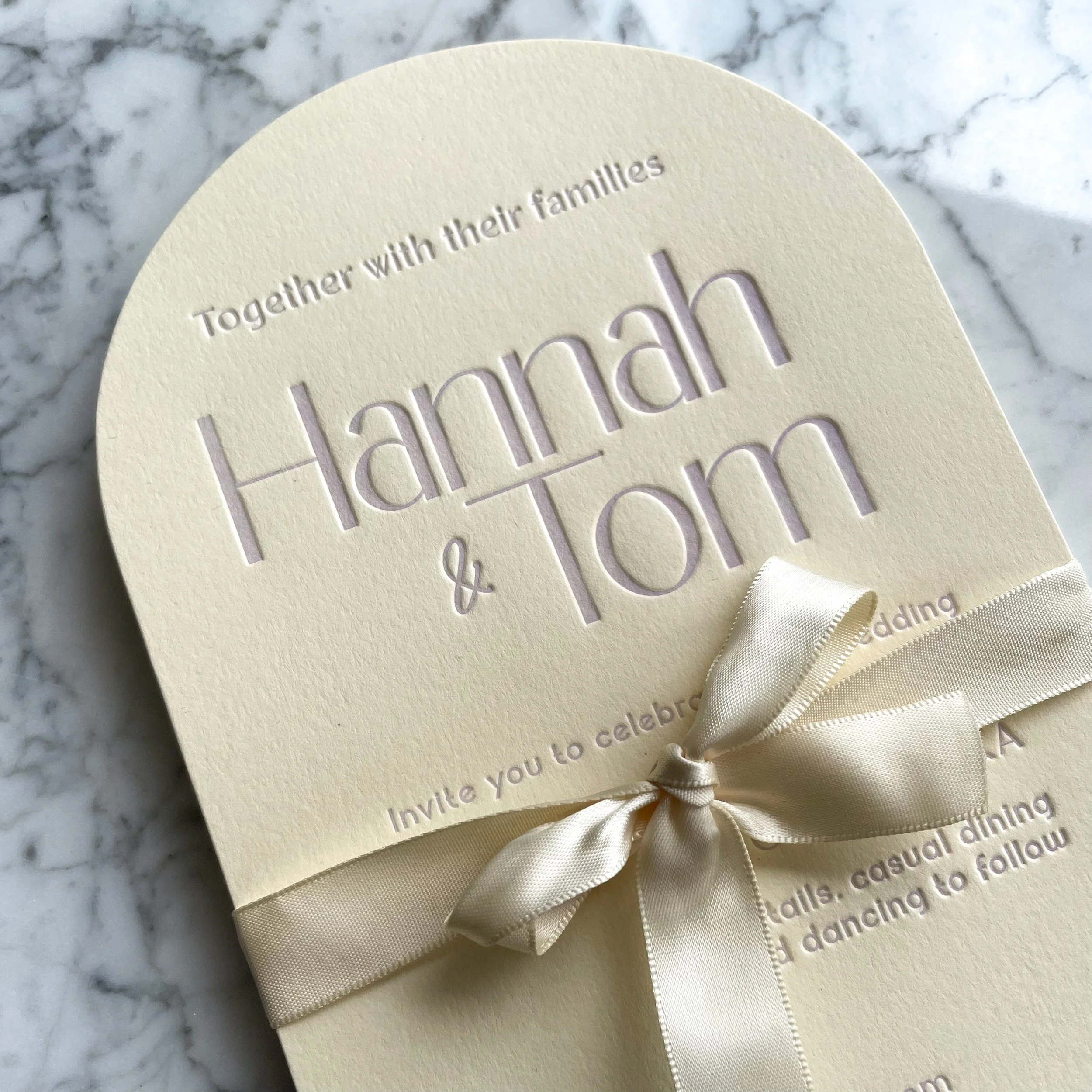 A close-up of a cream-colored wedding invitation with embossed text and a satin ribbon, placed on a marble surface.