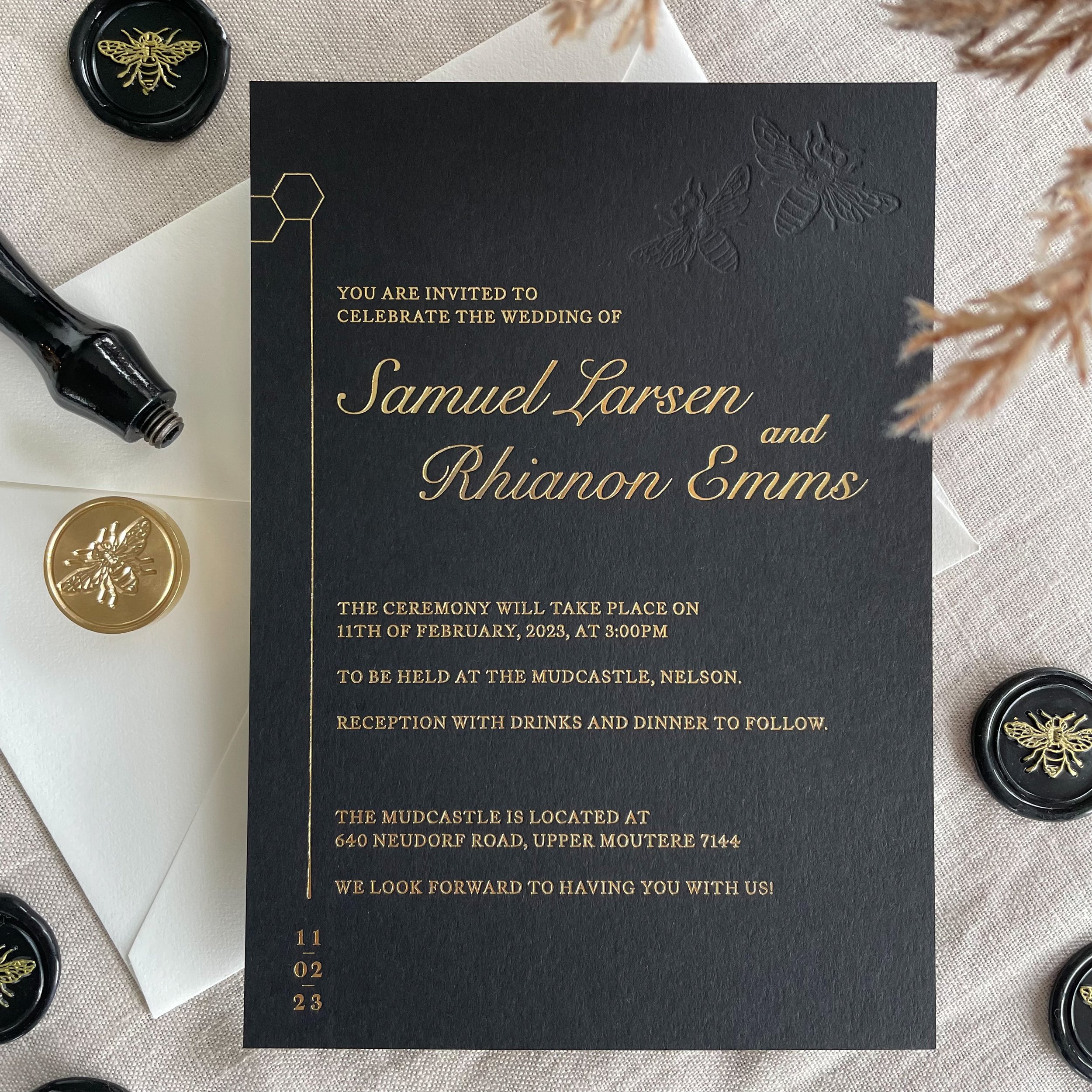 Wedding invitation card with gold embossed text and images of bees, surrounded by small bee stickers and a wax seal with a bee design, on a beige fabric surface.