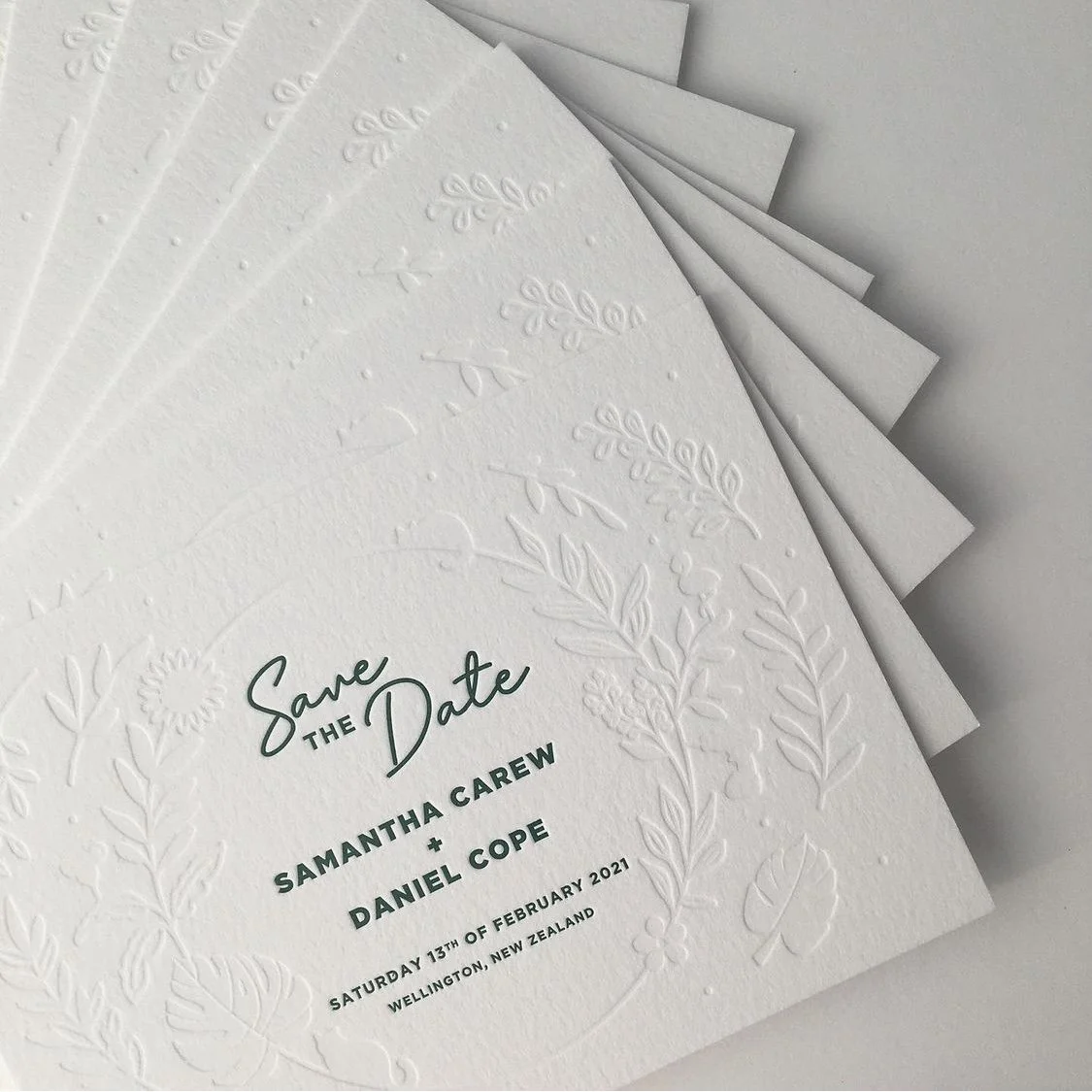 White wedding save the date card with embossed floral patterns, featuring text in dark green, including names, date, and location of the wedding.