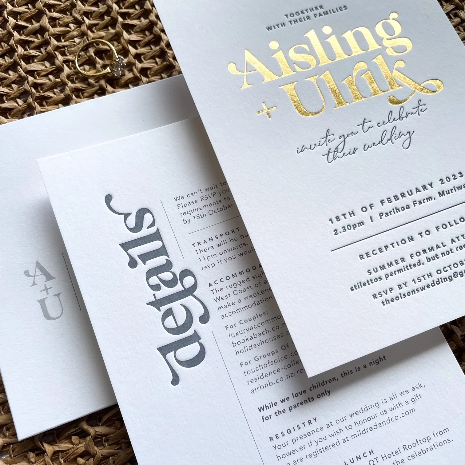 Invitation card for a wedding titled "Aisling + Uink." The card features gold foil text and includes event details such as the date, time, location, RSVP information, and a website. The wedding invitation is placed beside a small white booklet and a gold ring with a gemstone, all on a woven surface.