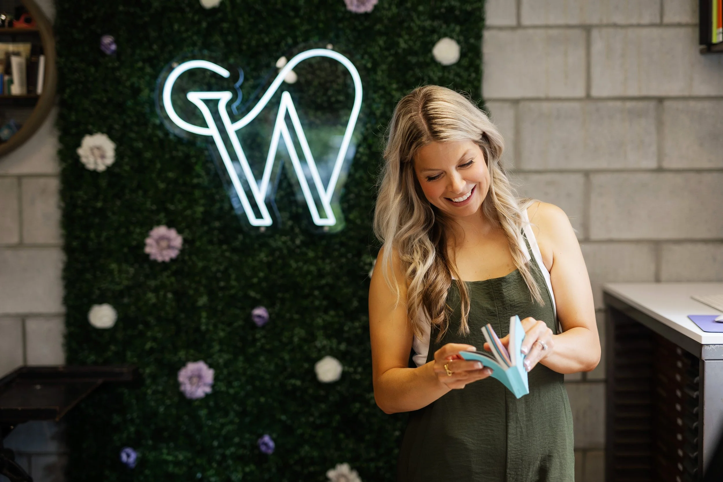 A smiling woman with long blonde hair wearing a green sleeveless jumpsuit, looking down at an open book she is holding, standing in front of a faux plant display on a brick wall with a neon 'W' sign.