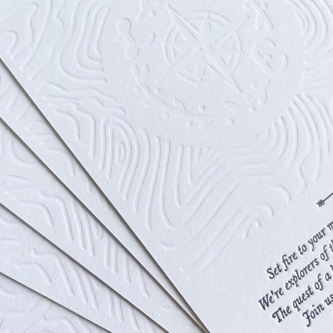 Close-up of white embossed paper with faint compass and map design, and partially visible text inviting explorers to join.