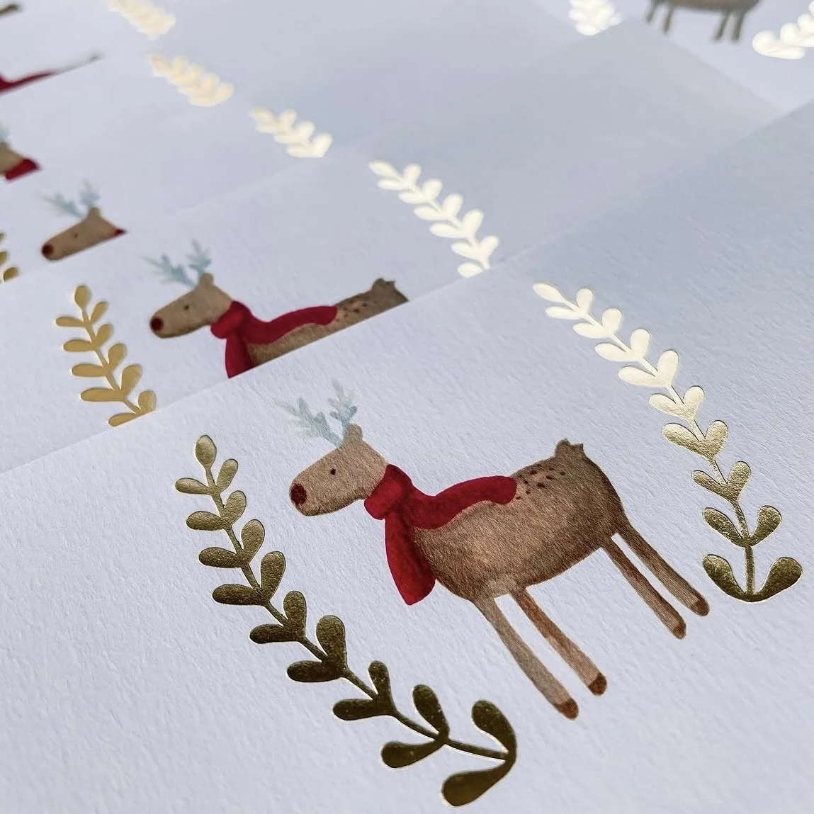 Holiday cards with illustrated reindeer wearing red scarves, surrounded by gold leaf designs on white textured paper.