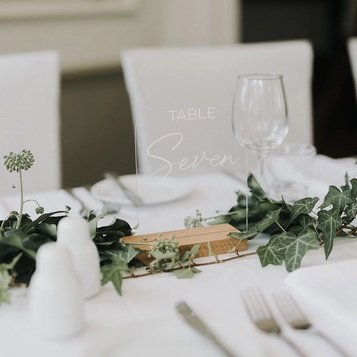 Elegant table setting with a clear acrylic sign displaying 'Table Seven', greenery, and white tableware.