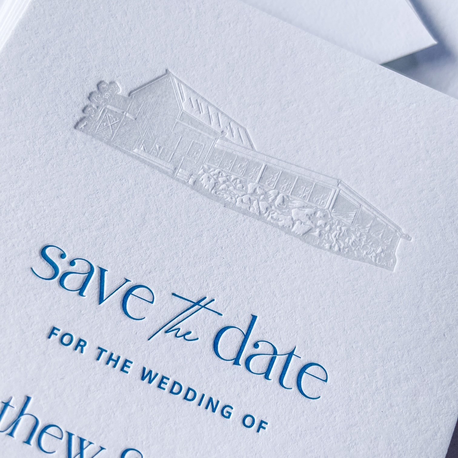 Close-up of a wedding invitation with embossed house illustration and blue text reading 'save the date'.