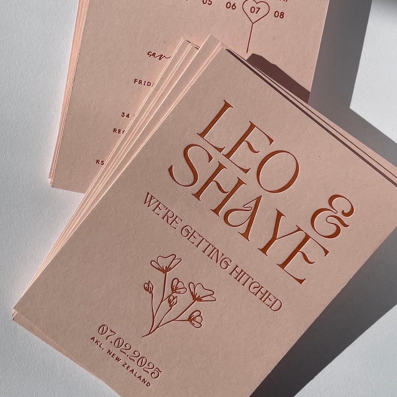 A wedding invitation card with pink background and elegant brown text that says "LEO & SHAYE" and "WE'RE GETTING HITCHED" with a floral illustration, date 07.02.2025, and location Auckland, New Zealand.