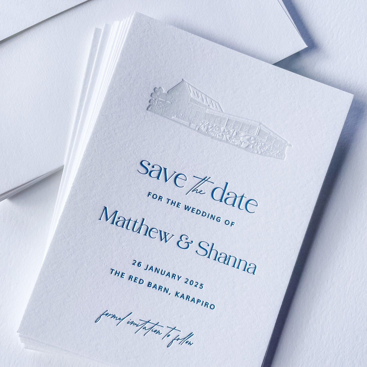 A close-up of a white wedding save-the-date card with embossed house and trees, featuring blue and black text for the wedding of Matthew and Shanna on January 26, 2025, at The Red Barn, Karpairo.
