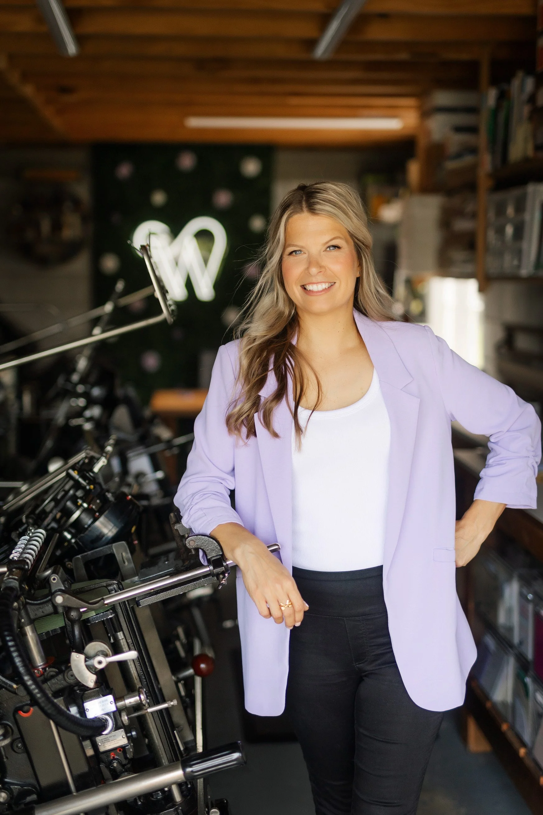 A woman with long wavy blonde hair wearing a lavender blazer, white top, and black pants standing in a workshop with letterpress printing machines, smiling at the camera.