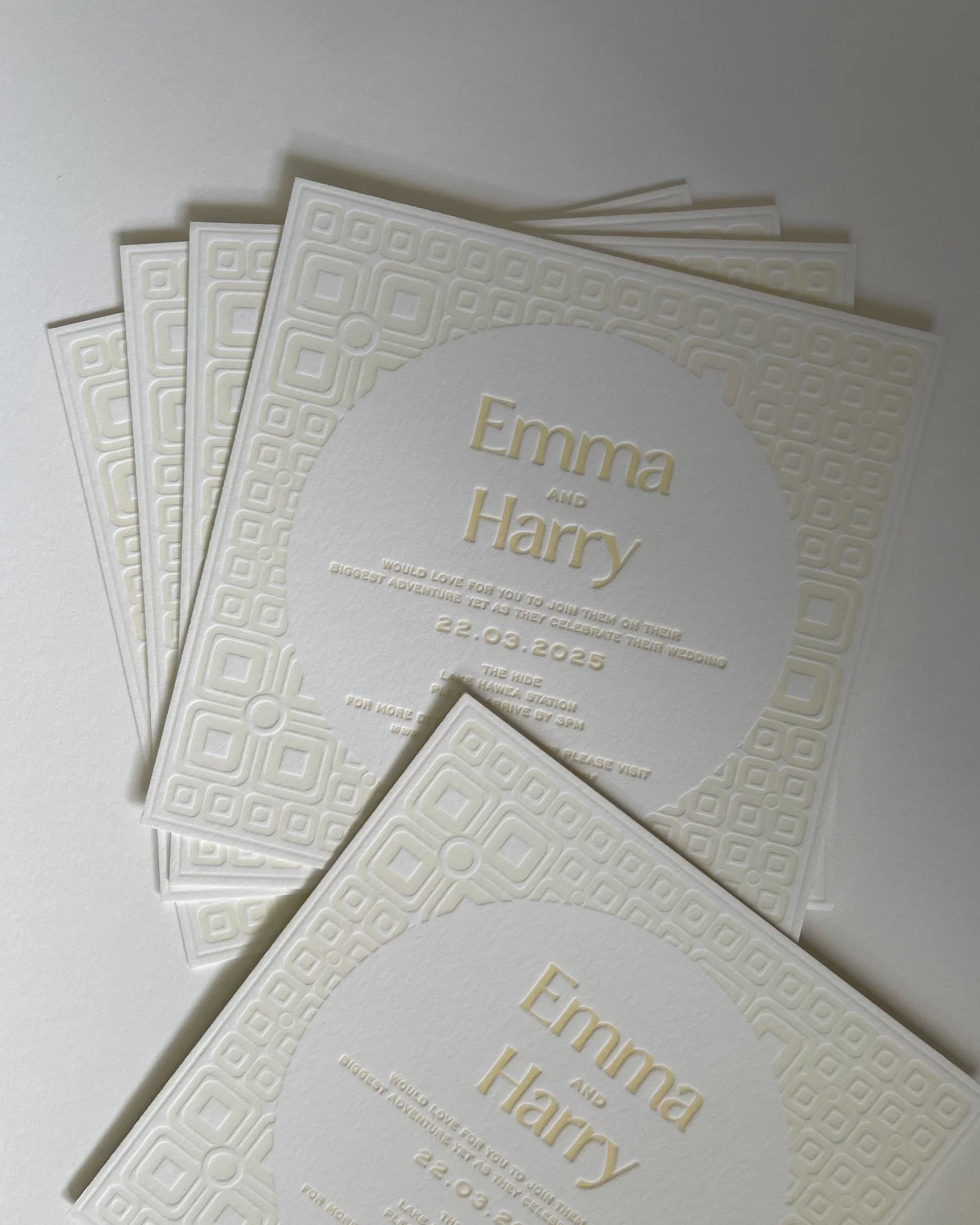 Close-up of white wedding invitation cards with intricate embossed geometric border and gold lettering for Emma and Harry's wedding.