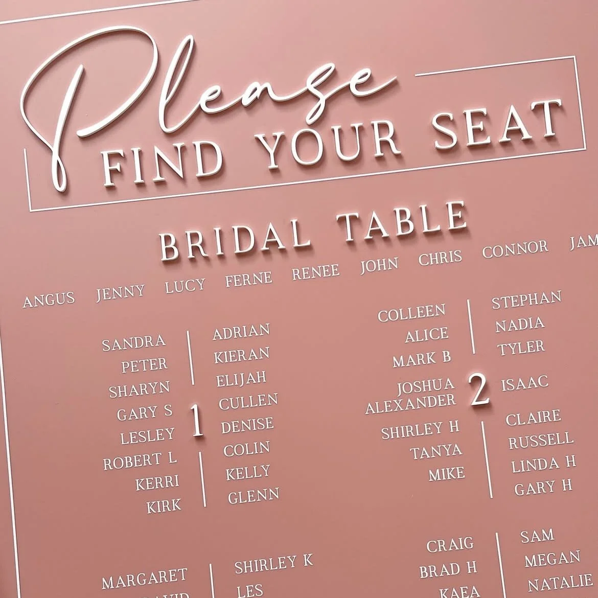A pink sign with white neon-style text asking guests to find their seat at a bridal table, listing guest names in columns under numbers 1 and 2.
