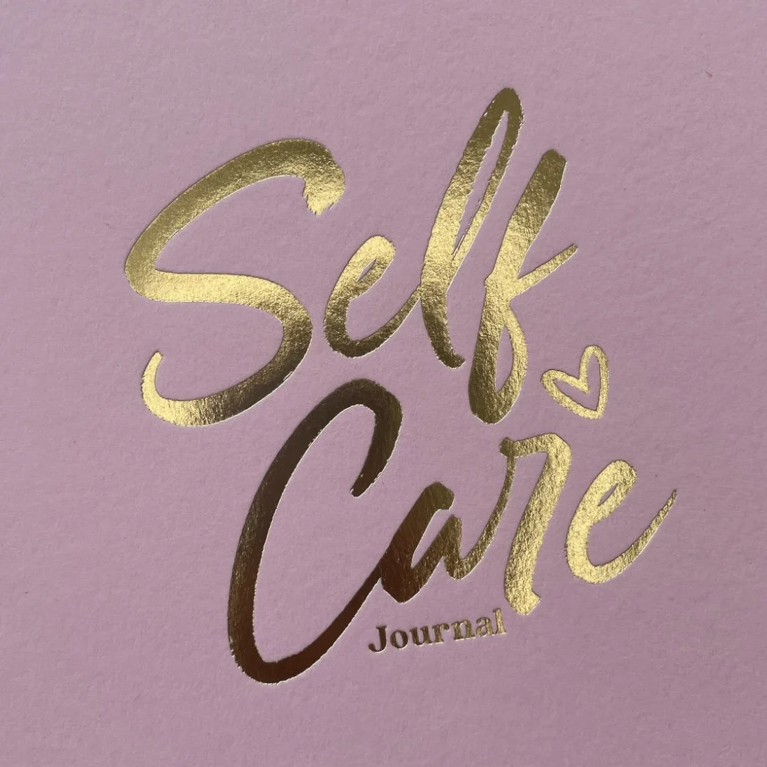 Pink journal cover with gold foil lettering that says 'Self Care Journal' and a small heart symbol.