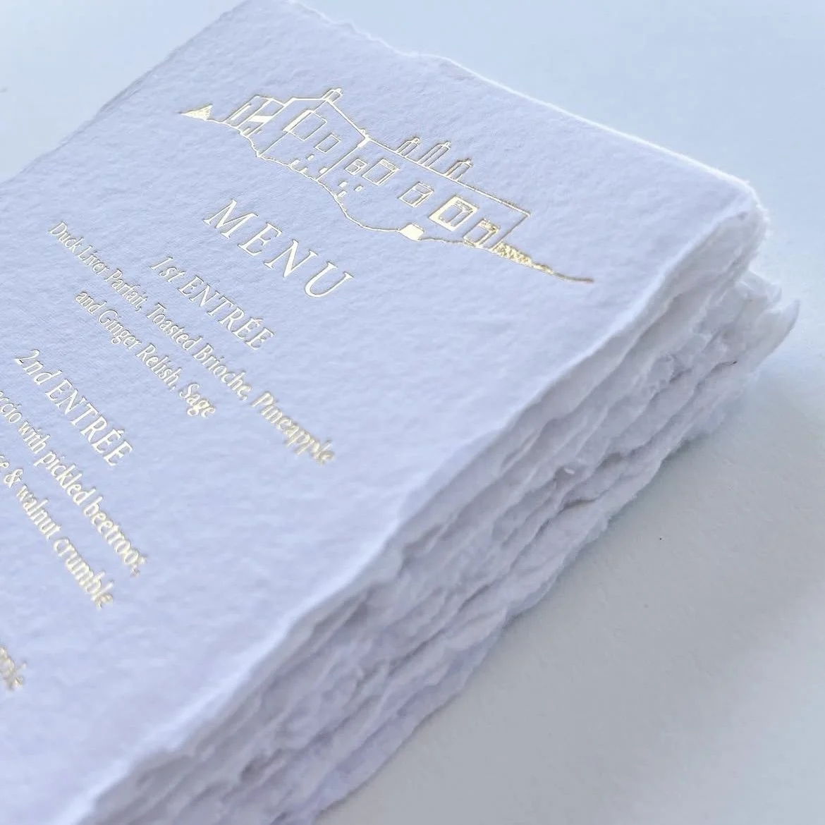A white textured paper menu with gold embossed house illustration and text, listing appetizers and entrees.