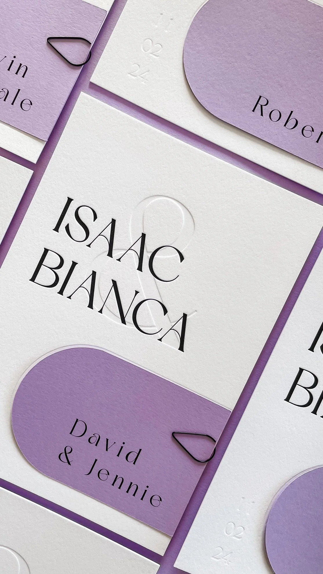 A collection of wedding invitation cards with a color scheme of white and purple, featuring elegant black and purple text with the names Isaac & Bianca, and additional names David & Jennie, with paper clips attached.